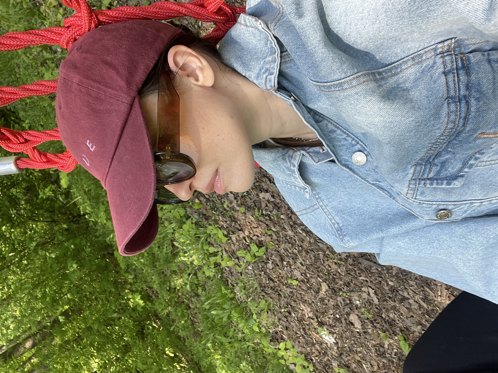 A person with light skin, wearing a denim jacket and a maroon baseball cap with white lettering, is seen lying in a red rope hammock in a park, possibly in Unknown, Unknown. Sunglasses rest on their face. The surrounding environment includes a mix of green grass and foliage, along with dry leaves and mulch on the ground.