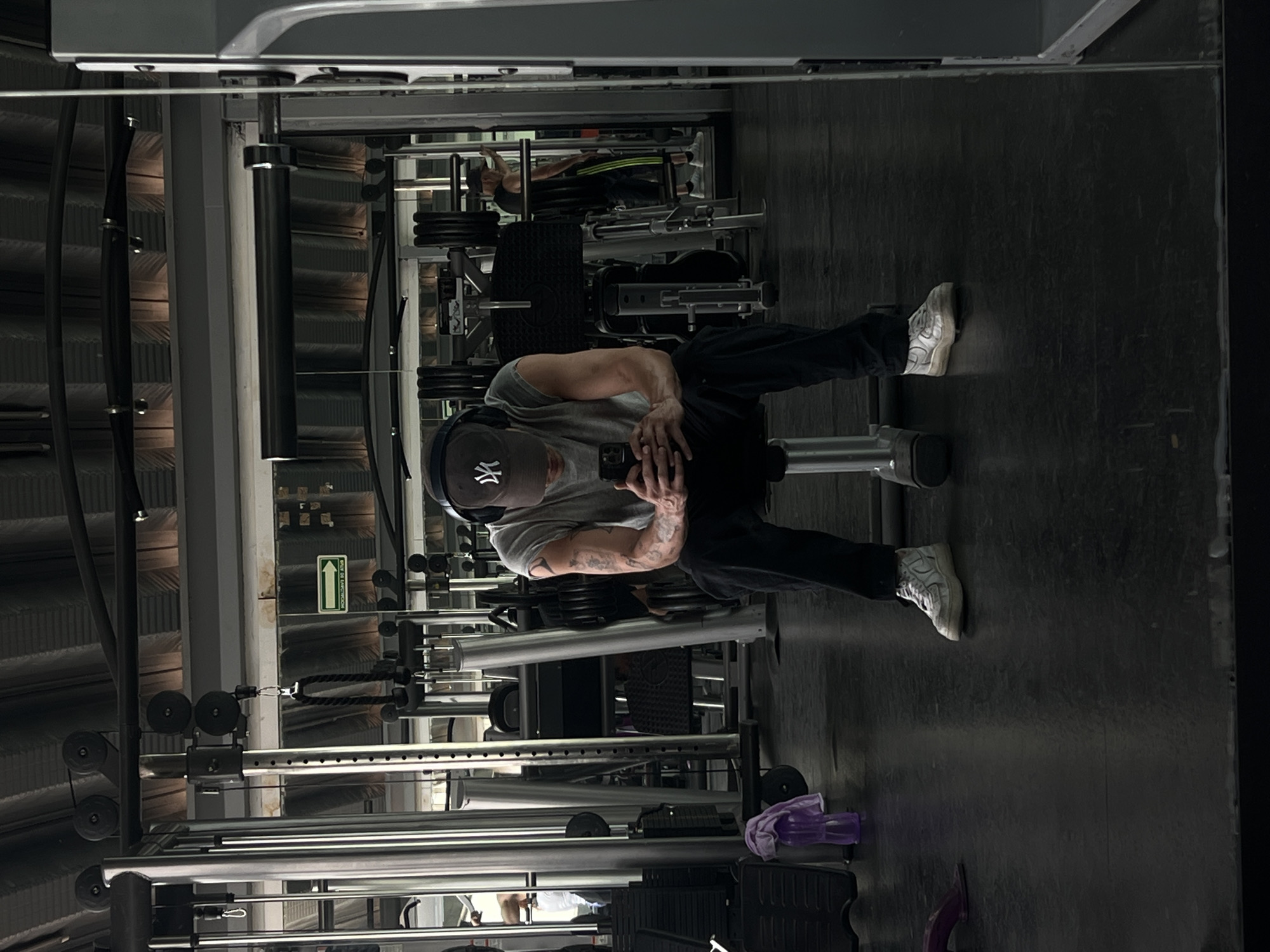 The image displays a man posing for a photo or selfie within a gym setting. He is positioned upright, wearing a grey t-shirt, dark pants, and white sneakers. A dark baseball cap with a white "NY" logo adorns his head, and headphones are draped around his neck. His muscular forearms, adorned with tattoos, are visible as he holds a smartphone with both hands, looking down at the device.
The background reveals typical gym equipment, including metal structures of cable machines, weight racks, and various exercise apparatus. Another individual can be faintly seen in the deeper background, utilizing a piece of gym equipment. A green "SALIDA DE EMERGENCIA" (Emergency Exit) sign, featuring an upward-pointing arrow, is mounted on a wall behind the man. Near his feet, a purple towel and a water bottle are visible on the dark gym floor. The indoor scene is illuminated by dim, artificial lighting. The overall impression is a moment of rest or a casual capture during a workout session in a gym located in Mexico City, Mexico.