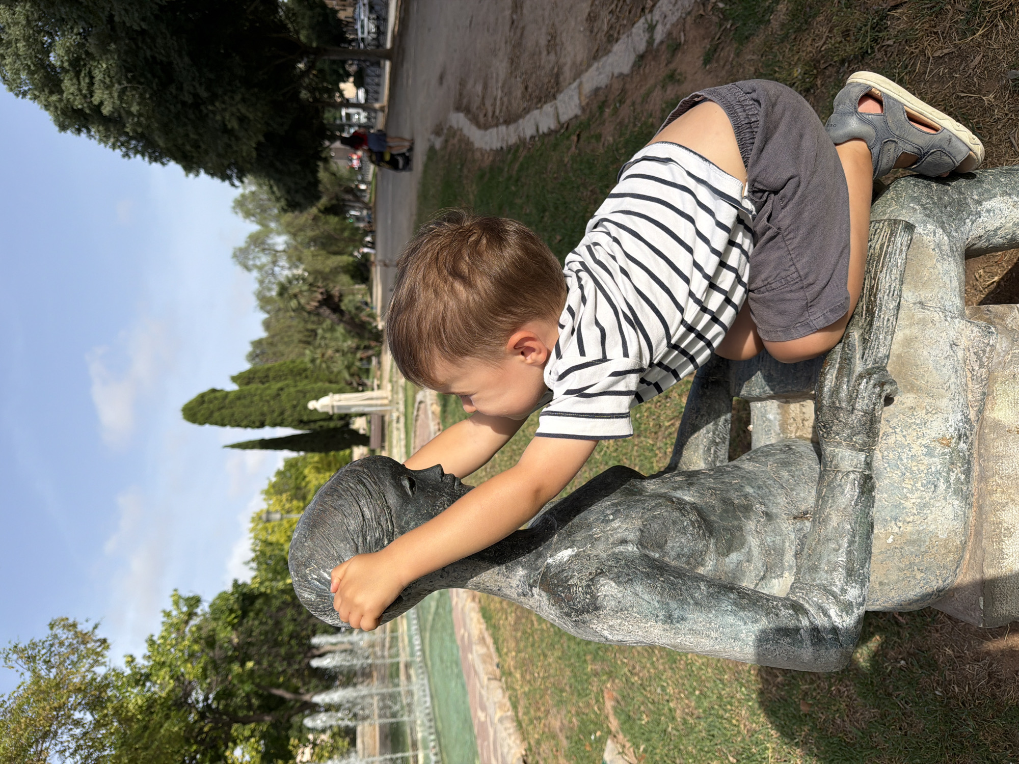 A young boy in a striped t-shirt and grey shorts kneels in front of a weathered bronze statue of a seated woman in a park. The boy has his hands on the statue's head, touching its hair. He appears to be a toddler, with light brown hair and fair skin. The statue is life-sized and seated on a stone bench. The background shows a park with trees, a fountain to the left, and a paved walkway with people and vehicles in the distance. The sky is blue with scattered clouds. The sun casts shadows on the ground, indicating it is daytime. The overall scene is peaceful and captures a moment of interaction between a child and art in an outdoor setting.
