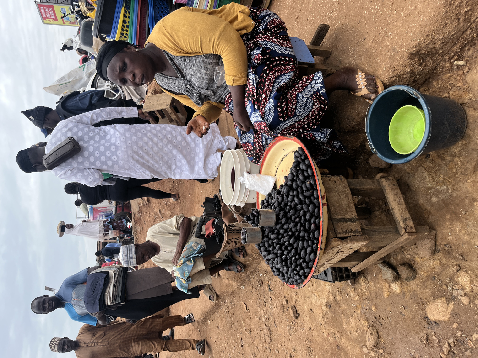 The image depicts a market scene in Jos, Nigeria, under a cloudy sky. In the foreground, a woman wearing a yellow cardigan and a blue, red, and white patterned skirt sits on a blue stool. She is looking towards the left of the frame with her mouth slightly open and her right hand is raised, holding what appears to be a small white object, possibly food or a coin. In front of her is a large, round tray filled with numerous dark, oval-shaped fruits, likely olives or a similar local produce. A smaller metal can is stacked on top of another, also containing some of these fruits. Next to the tray is a white bucket containing a dark liquid. To the woman's left, a man in a light-colored tunic and a white and blue cap is seated, appearing to be working with some fabric and a blue and white cloth. Further back, several other people are visible. A man in a brown tunic and a striped cap stands on the left, holding a stack of folded garments. Another man in a blue shirt and cap stands next to him, also holding folded clothing. In the background, a person in a white traditional outfit with a patterned design walks away from the camera. The ground is predominantly a reddish-brown dirt, with some scattered stones and patches of dampness, suggesting it might be a roadside or unpaved market area. In the far background, there are glimpses of stalls, vehicles, and more people, indicating a busy market environment. A sign with text in an unfamiliar script is visible in the upper right portion of the image.