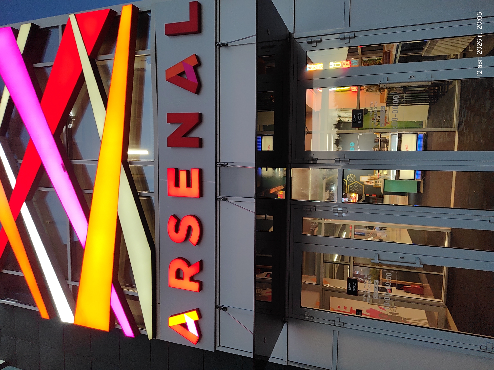 The image shows the exterior of a building with a prominent red sign spelling "ARSENAL" in large, illuminated letters. The building's facade is decorated with abstract, multi-colored light strips in shades of orange, pink, and white, creating a modern and vibrant aesthetic. Below the sign, there are glass doors leading into the building, reflecting the surrounding environment. A sign on the door reads "КИРУ ВХОД" (KIRU VKHOD), which translates to "Entrance." Another sign indicates operating hours as "0:00-0:00". A timestamp in the bottom right corner of the image reads "12 авг. 2026 г., 20:05". The lighting suggests it is either dusk or evening. The location is identified as Astana, Kazakhstan.