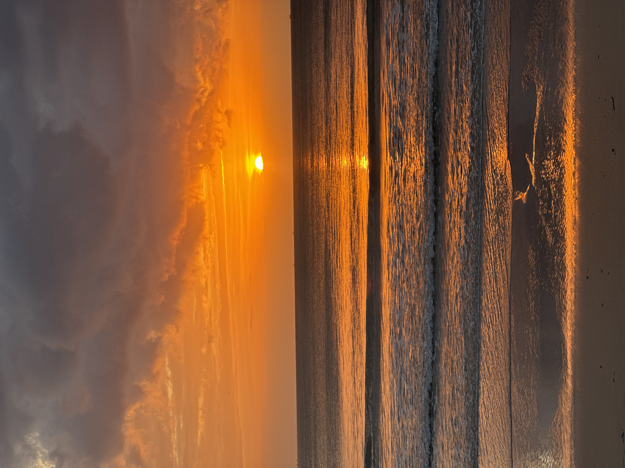 The image captures a serene sunset over the ocean in Kuta Selatan, Indonesia. The sky is ablaze with vibrant orange and yellow hues, with streaks of dark gray clouds adding dramatic contrast. The sun, a glowing orb, is partially visible, casting a shimmering golden path across the calm water. Gentle waves roll towards a sandy shore, their crests reflecting the warm colors of the sky. The beach is mostly devoid of people, with only a few dark specks suggesting scattered debris or footprints. The overall impression is one of tranquility and natural beauty.