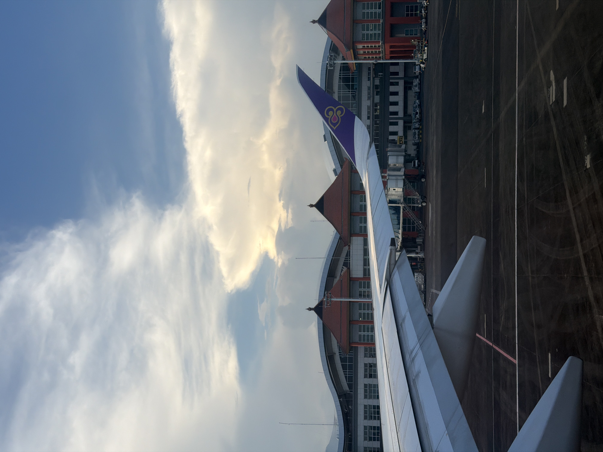 This image depicts an airplane wing with a tail fin bearing the Thai Airways International logo, seen through the window of the aircraft at Ngurah Rai International Airport (DPS) in Kuta, Bali, Indonesia. The airplane is on the ground, parked at the gate. The view includes part of the airport terminal building, the taxiway and the sky with clouds. The overall lighting appears to be daylight.