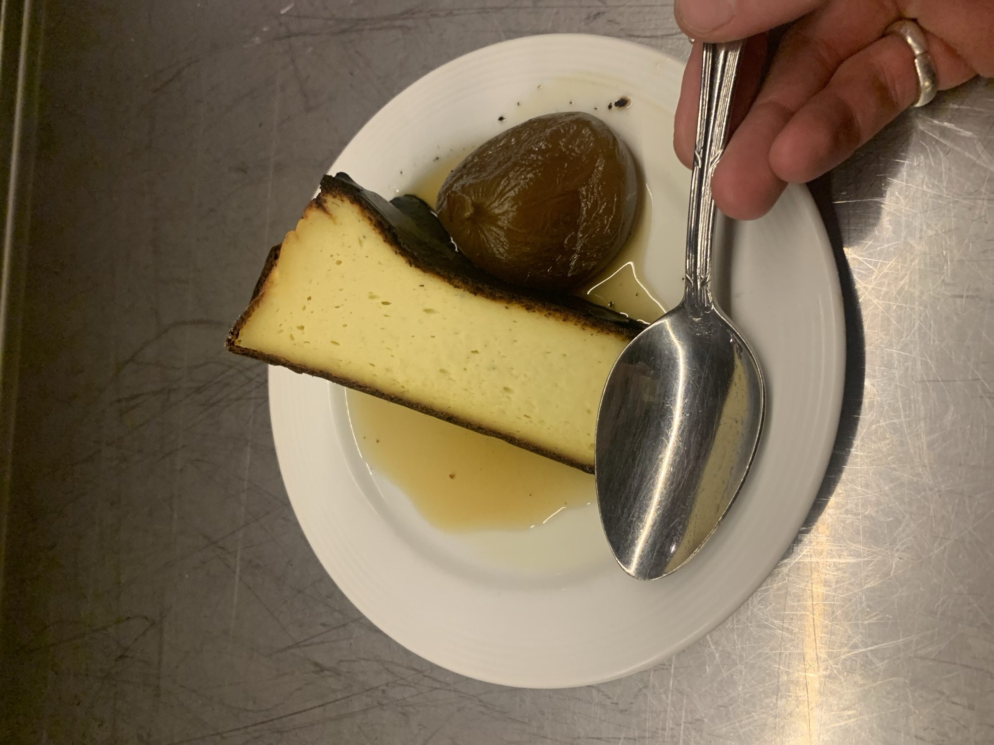 A triangular slice of burnt Basque cheesecake and a poached fig are on a white plate, which rests on a stainless steel surface. A hand holding an ornate silver spoon appears to be about to take a bite or spread the sauce. The location is likely a restaurant in Buenos Aires, Argentina, based on the style of dessert, but no defining landmarks are visible.