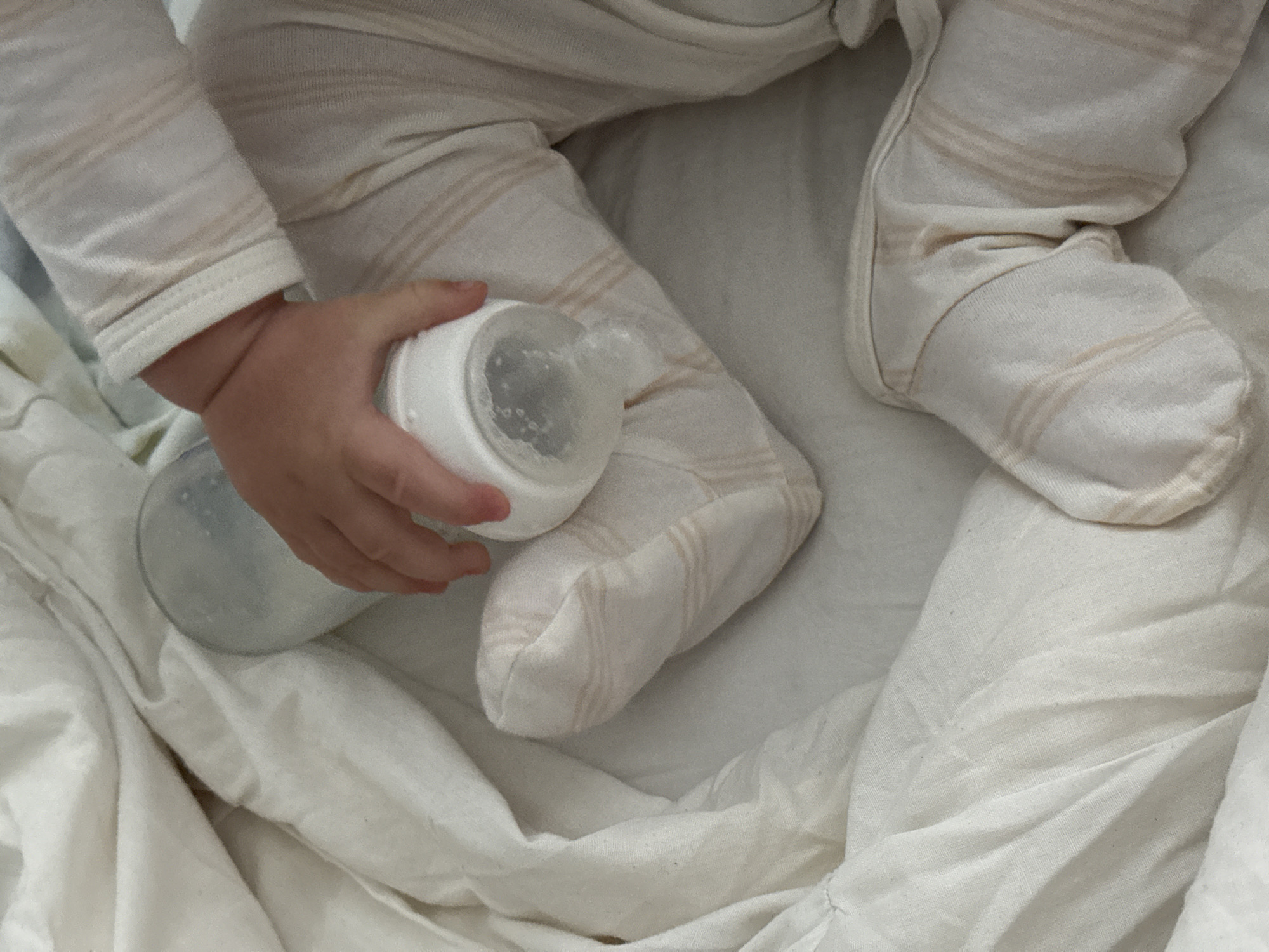 A close-up, overhead shot focuses on a baby's hand holding a partially filled baby bottle amidst crumpled white bedding. The baby is wearing a cream-colored footed pajama set with subtle light brown stripes. The bottle, made of clear plastic with a white nipple and cap, is angled, revealing a small amount of liquid inside. The background is dominated by the texture and folds of the white bedding, which appears to be a crib or bassinet. The lighting is soft and diffused. The image does not provide any specific information that would allow for location within the city of Siegburg, Germany.