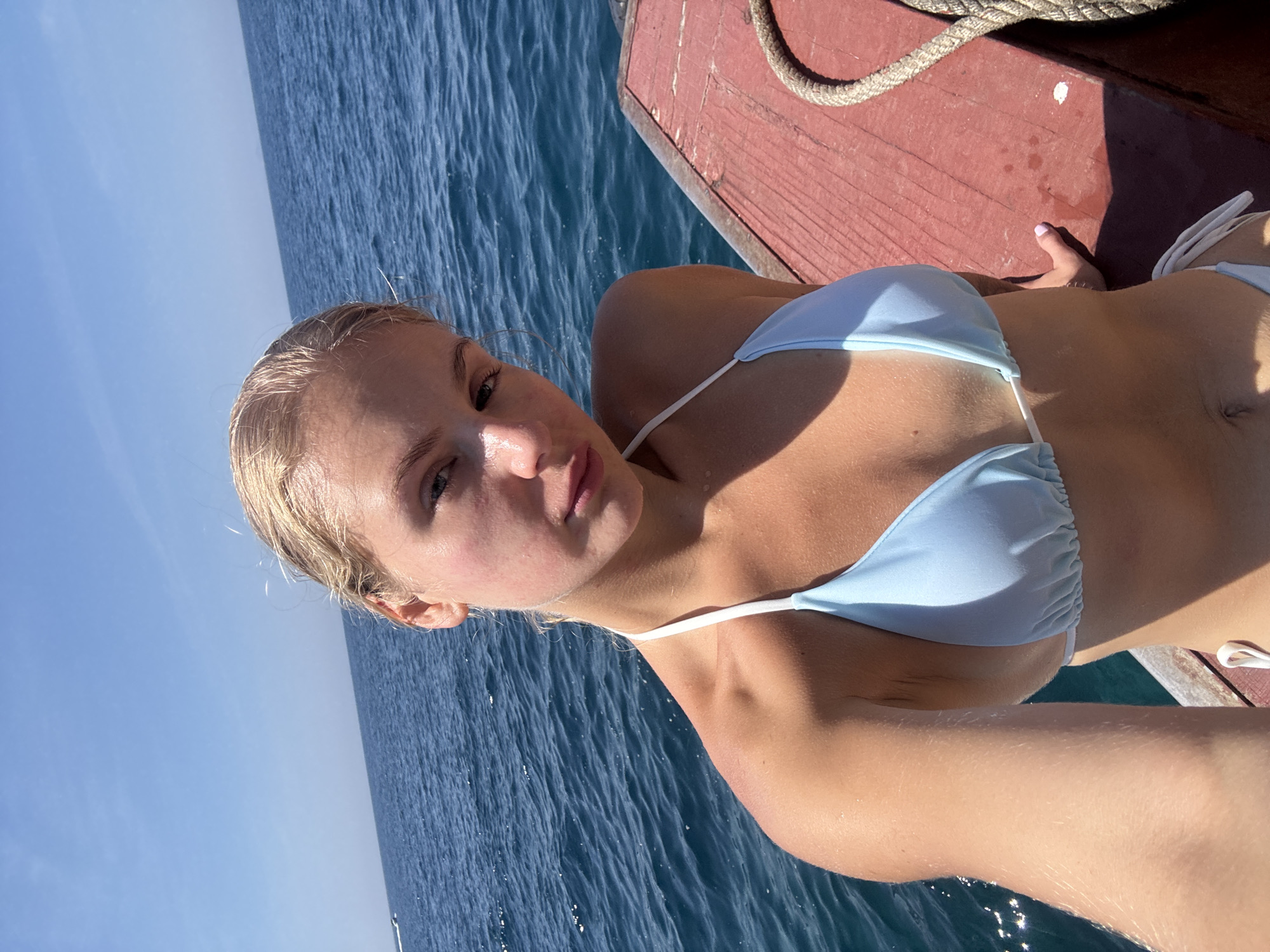 A young woman with blonde hair tied back is taking a selfie while sitting on the edge of a wooden boat. She is wearing a light blue bikini and her left arm is extended towards the camera. The background shows the blue ocean and a clear sky. The wooden surface of the boat appears worn and reddish-brown, and a thick rope is visible to the right. The overall scene suggests a sunny day out on the water. There is no indication in the image that this location is in Nha Trang, Vietnam.