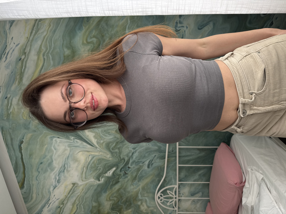 A female subject stands in what appears to be a bedroom in Scafati, Italy. She is positioned slightly angled, facing the camera with a subtle smile. The subject has long, brown hair and wears round eyeglasses with dark frames. She is dressed in a short-sleeved, grey, ribbed crop top and light beige corduroy trousers. The wall behind her features a marbled pattern in shades of green, turquoise, and off-white. To the left, a white metal bed frame with a decorative headboard is partially visible, holding a pink pillow on white bedding. A white curtain or window covering is present on the right side of the frame.