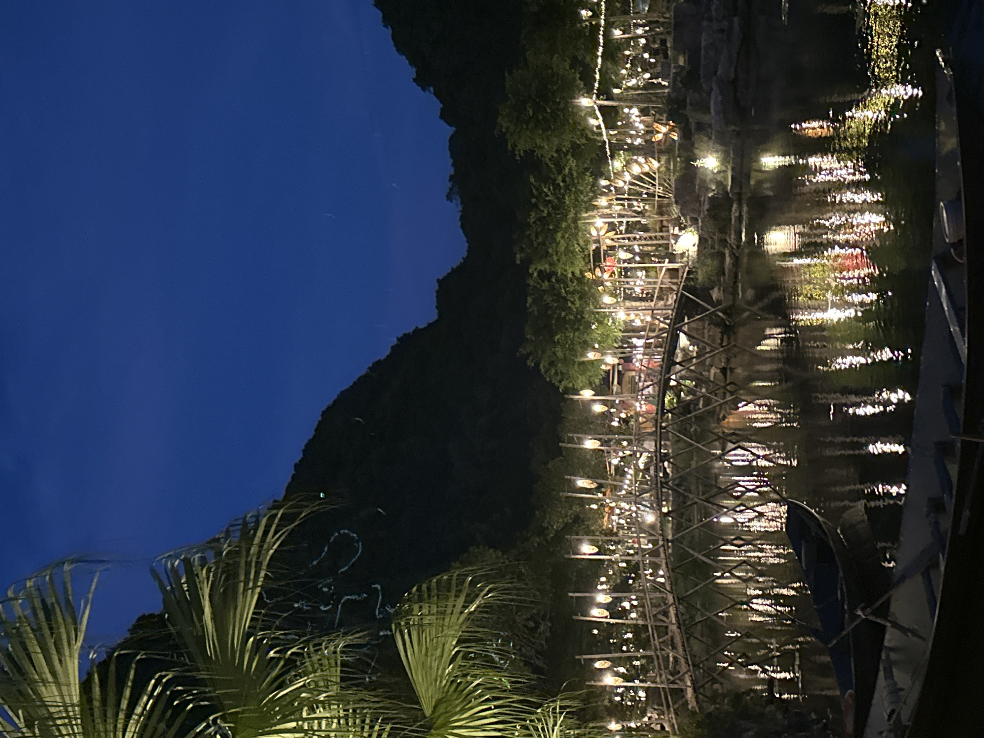 In the city of Hoa Lư Ward, Vietnam, at dusk, a tranquil scene unfolds by a body of water. A rustic wooden bridge, adorned with numerous hanging lanterns, arches gracefully over the water. The lanterns cast a warm, inviting glow, their reflections shimmering on the dark surface of the water below. Trees, illuminated by the soft light, line the banks of the water, creating a festive atmosphere. The background is dominated by dark, silhouetted mountains, against a deep blue sky. In the foreground, two boats are moored, their forms barely visible in the dim light, adding to the peaceful ambiance of the night.