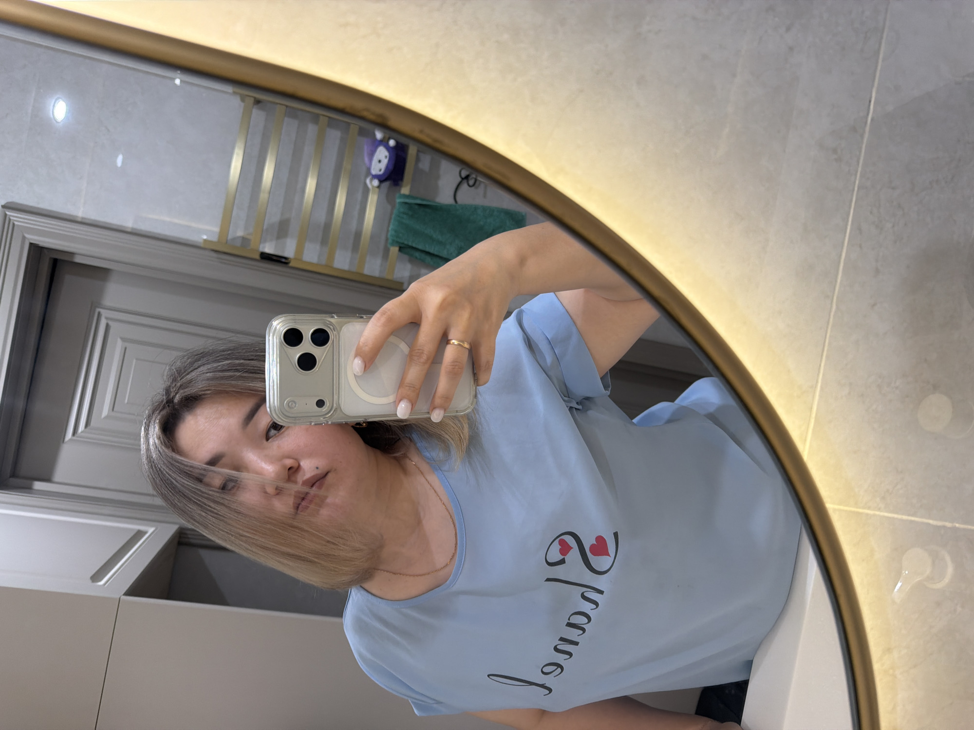 A woman is taking a mirror selfie in a modern bathroom. She is wearing a light blue t-shirt with black text and two red hearts. Her hair is blonde and cut in a bob style. She is holding a smartphone in her right hand, which is capturing her reflection. The bathroom features a large, oval mirror with a golden frame and integrated lighting. In the background, a towel rack with gold bars is visible, along with a green towel and a small purple toy. The walls are tiled with a light grey marble pattern. The overall aesthetic suggests a clean and contemporary living space, likely in a residential building in Astana, Kazakhstan, given the context provided.