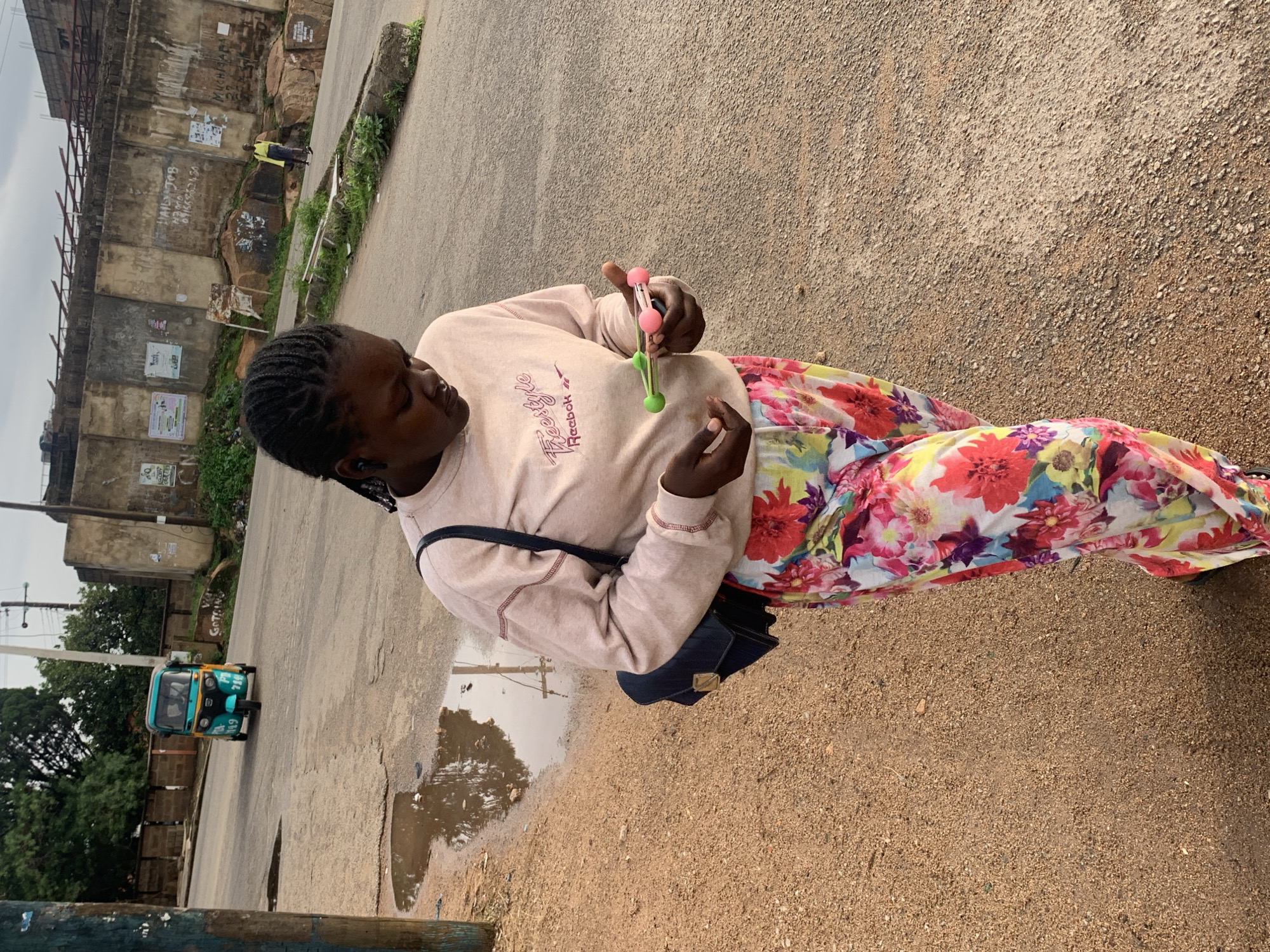 A young Black girl with braided hair is standing on a dirt and asphalt road, holding a colorful toy. She is wearing a light pink long-sleeved shirt with a graphic design and floral pants. A black purse is slung across her chest. The scene appears to be in Jos, Nigeria, with a building and some greenery visible in the background. A blue and yellow auto-rickshaw is driving down the road. There are puddles of water on the ground, suggesting it may have recently rained. The sky is overcast.