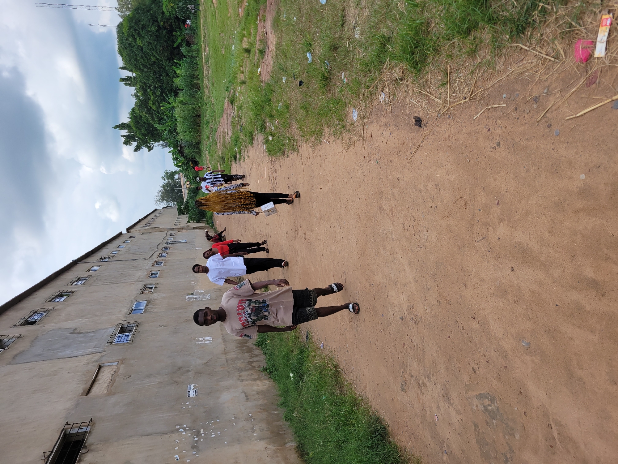 The image depicts an outdoor scene in Egbagu, Nigeria. A group of individuals are walking along a dirt path beside a long, weathered building with numerous windows, some barred. To the right of the path, there is a grassy area with some vegetation and debris, and in the background, a dense line of trees. The sky is overcast with clouds. In the foreground, a young man wearing a light brown t-shirt with a graphic print and dark shorts walks towards the camera. Behind him, a person in a white garment and black trousers is walking away, followed by a person in a red top and black trousers. Further along the path, a person with long blonde braids is walking away, carrying a bag. In the distance, other figures can be seen. The overall atmosphere is casual, with people moving along the path in a rural or semi-urban environment.