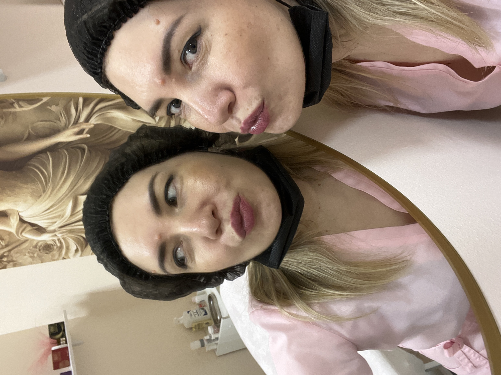 The image shows a woman taking a selfie in a mirror. She is wearing a black hair net and a pink robe, with a black mask partially covering her face. The reflection in the mirror shows her face in close-up, with makeup on her eyes and lips. Behind her, a portion of a large framed artwork with a classical depiction of a woman is visible. The setting appears to be an indoor space, possibly a salon or a spa, given the presence of what look like beauty products on a shelf in the background. The lighting is soft and even, suggesting an indoor environment. The woman's expression is somewhat playful, with her lips pursed. There is no visible text in the image. The location context of Sochi, Russia, does not provide any specific visual cues within the image itself.