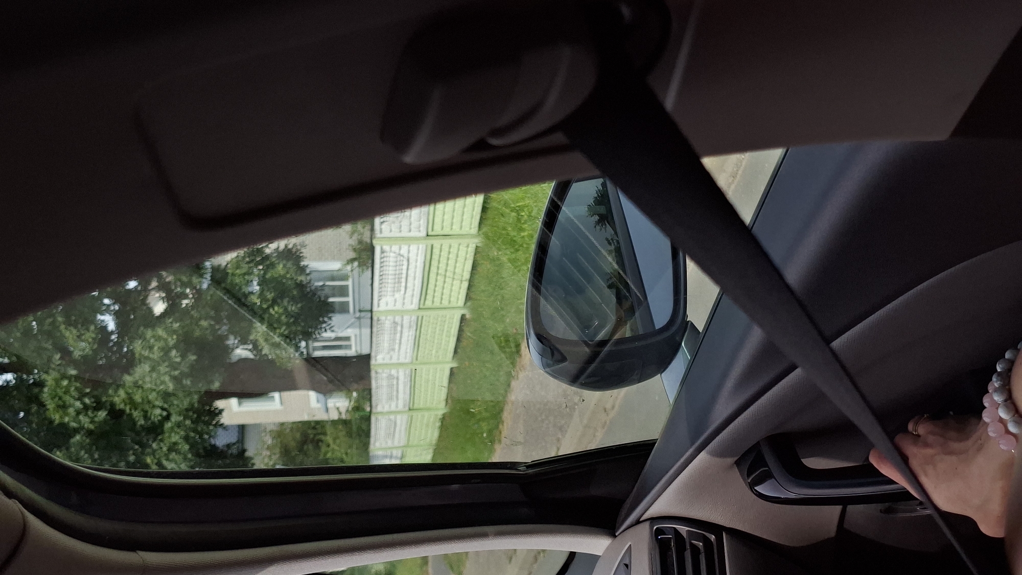 The image is a first-person perspective from inside a vehicle, looking out the passenger side window. A black seatbelt is prominently positioned across the foreground, running diagonally from the upper right to the lower left. In the lower left corner, a bare foot with a ring on one toe and beaded bracelets on the ankle is visible, resting near the door handle.
Through the window, a street is visible, bordered by a light green and white picket fence. Beyond the fence, there is a grassy area and then a building with white window frames. A large tree with green foliage stands in front of the building. A side-view mirror is visible, reflecting the sky and trees.
The scene appears to be on a street in Myadzyel, Belarus.