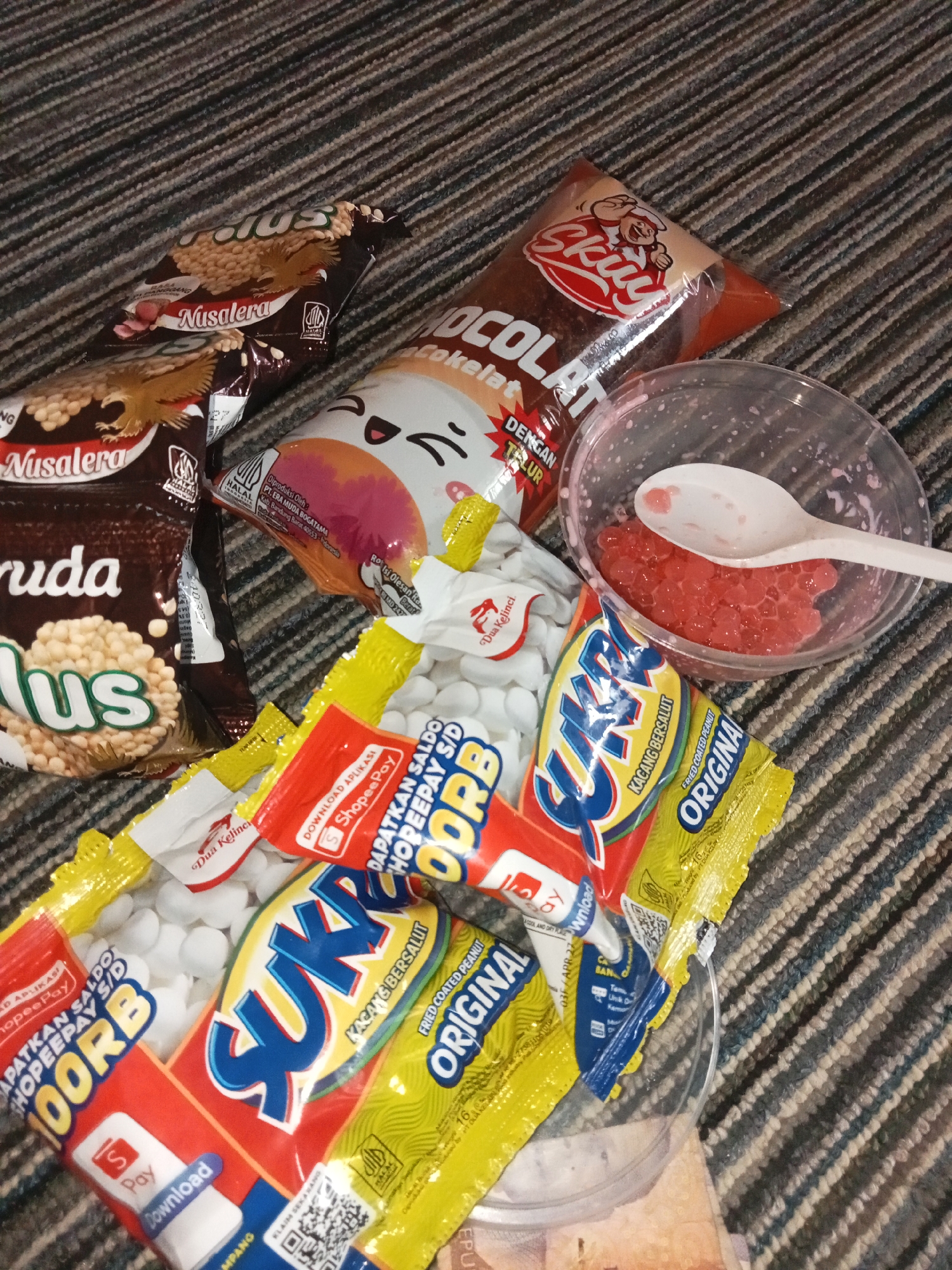 A close-up, overhead shot shows a variety of snacks and a small clear cup containing red boba pearls and a white spoon, arranged on a textured, striped carpet. The snacks include two bags of "Nusarasa" peanuts, one labeled "Plus," and a packet of "Skuy Chocolate" with a cartoon drawing of a chef. Several yellow and red packaged snacks with the brand name "Suk" are visible, with text indicating "KACANG BERSALUT" and "ORIGINAL." One red packet also shows a ShopeePay logo and a QR code. The scene is dimly lit, suggesting it was taken indoors. No people are present, and the setting does not provide specific information about the city of Pancurwening, Indonesia.