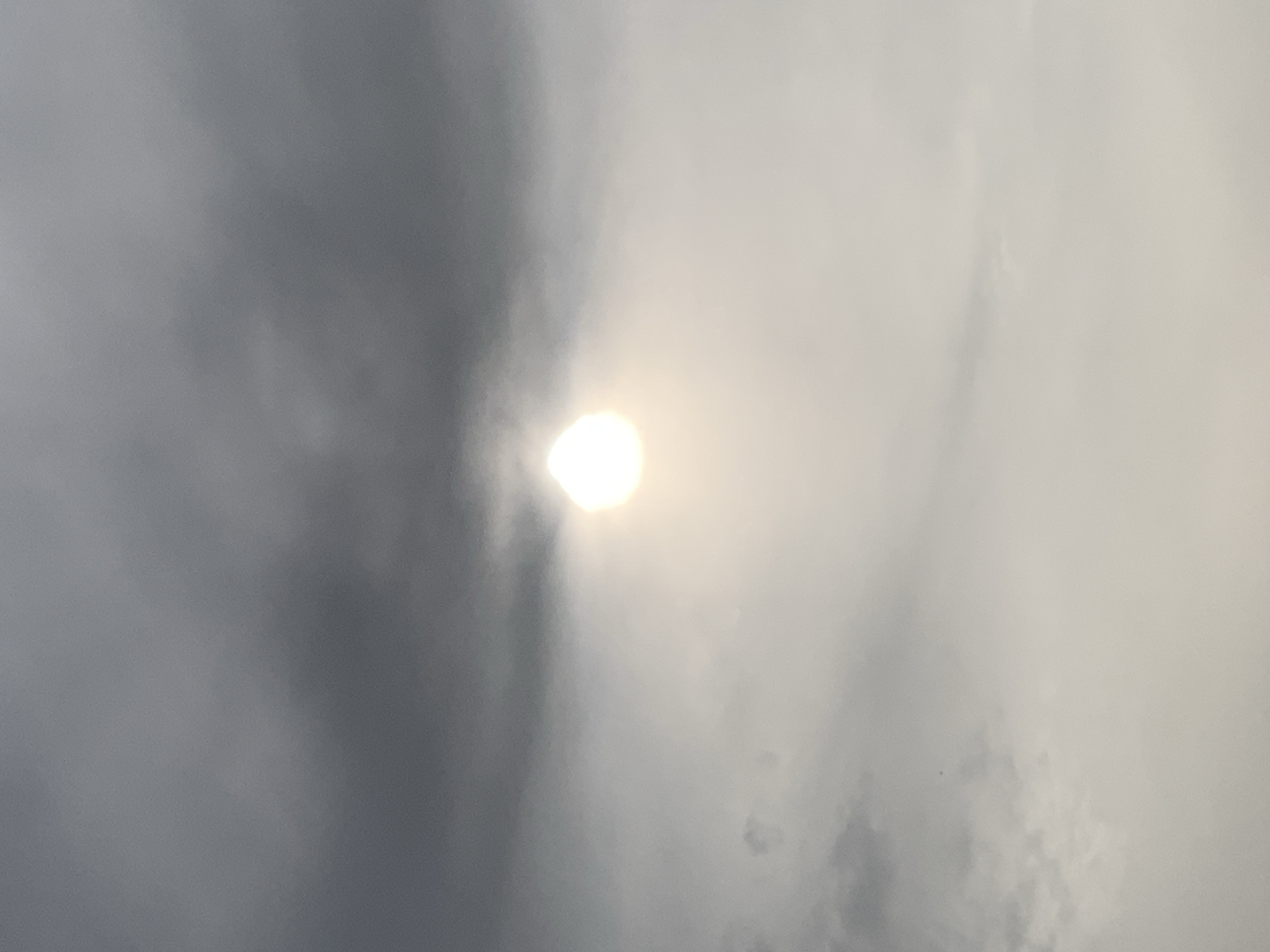 The image shows a bright sun obscured by thick clouds, likely in Adankolo, Nigeria. The sky ranges from dark gray on the left side to light gray/white on the right, indicating varying cloud density. No people, objects, or actions are visible other than the diffused sunlight through the clouds.