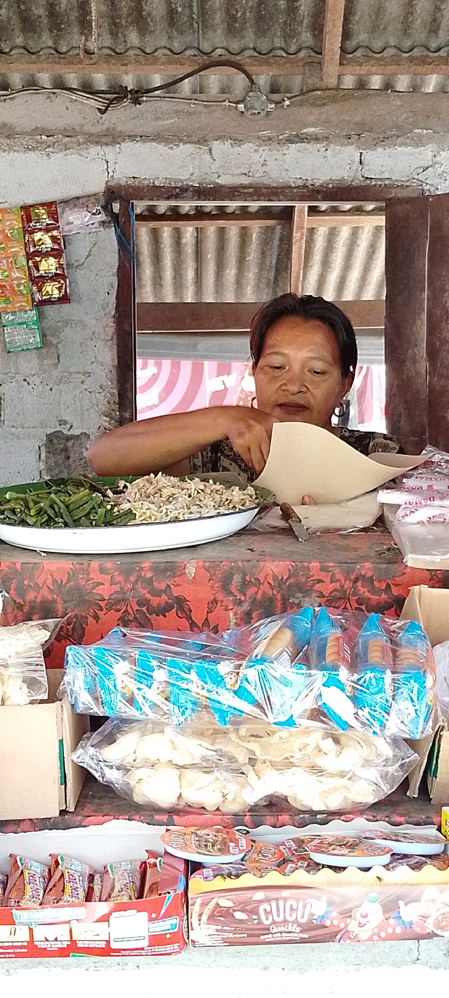 A woman of Asian descent is seen behind a counter in an outdoor market stall in Pekutatan, Indonesia. She is wearing a dark patterned top and hoop earrings. The woman is in the process of wrapping something in a light brown paper, her hands and forearms are visible. In front of her, on the counter, is a white oval dish containing a variety of food items, including what appears to be chopped green beans and a pile of small, light-colored shredded food. The counter is covered with a red cloth featuring a dark floral pattern.
Behind the woman, the stall is constructed with rough, white-painted cinder blocks and has a corrugated metal roof supported by wooden beams. Electrical wiring is visible along the ceiling. To the left of the woman, several small, colorful packets, possibly of snacks or spices, are hanging on the wall.
Below the counter, the scene is filled with various packaged goods. In the foreground, there are several rows of packaged snacks. Some are in clear plastic wrapping and contain cone-shaped items, possibly cookies or pastries, with blue and yellow packaging. Below these, more packaged snacks are visible, including what looks like crackers or chips. To the far right, a cardboard box contains more packaged goods, including a large chocolate bar with the brand name "CUCU" prominently displayed. The overall impression is of a small, local food stall operating in an open-air market.