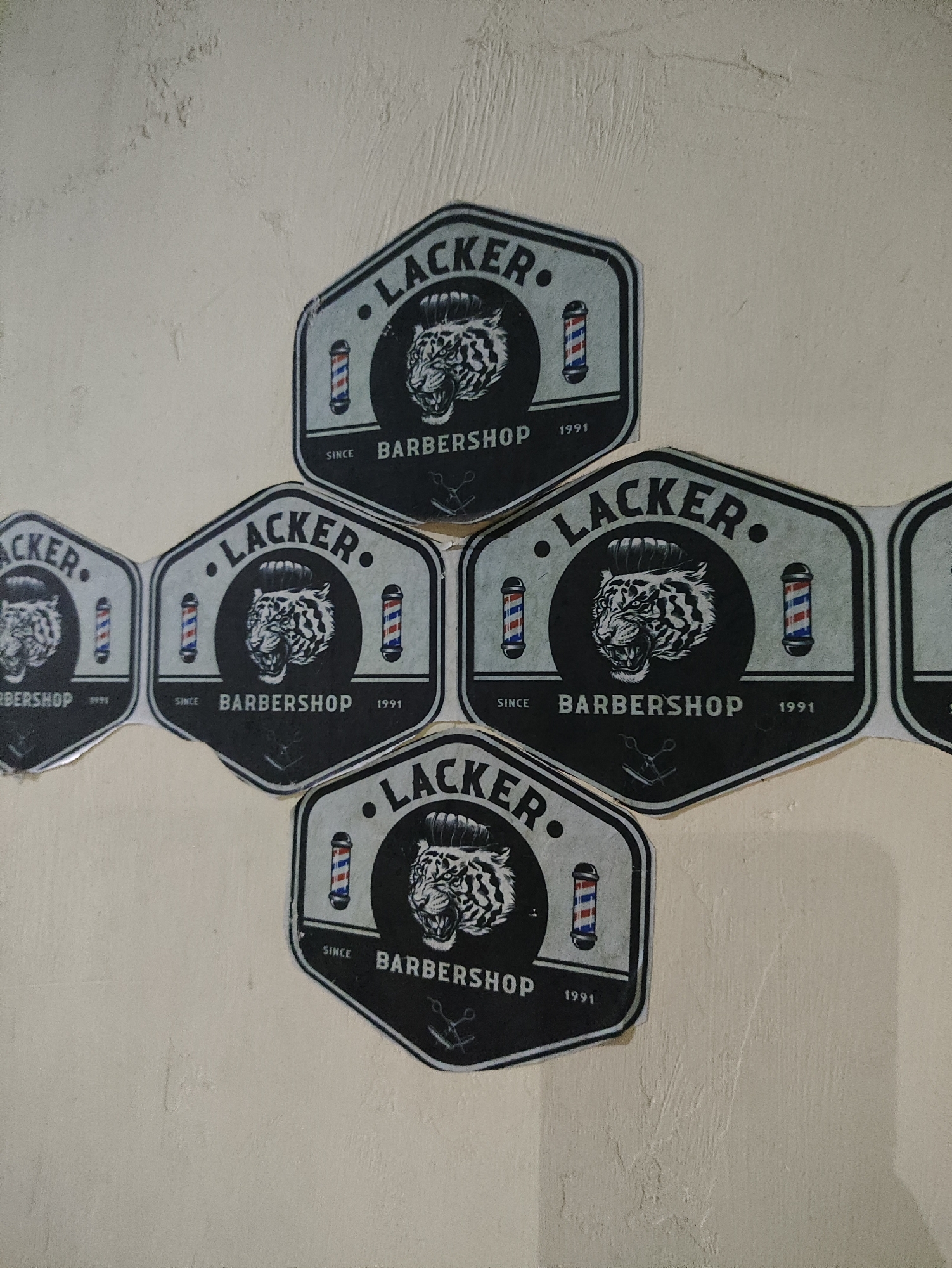 This image displays a collection of circular stickers arranged in a diagonal, diamond-like pattern on a textured, light-colored wall. The stickers are silver-bordered with a black background, each featuring a stylized tiger's head with an elaborate hairstyle, accompanied by barber poles on either side. Text on each sticker reads ".LACKER. BARBERSHOP SINCE 1991". The stickers appear to be uniform in design and are placed in close proximity, overlapping slightly in some areas. The wall surface shows signs of wear and has a rough texture, typical of an interior wall. There are no people or animals present in the image. The scene is static and focuses solely on the decorative stickers on the wall. This image was captured in the city of Kepung, Indonesia.