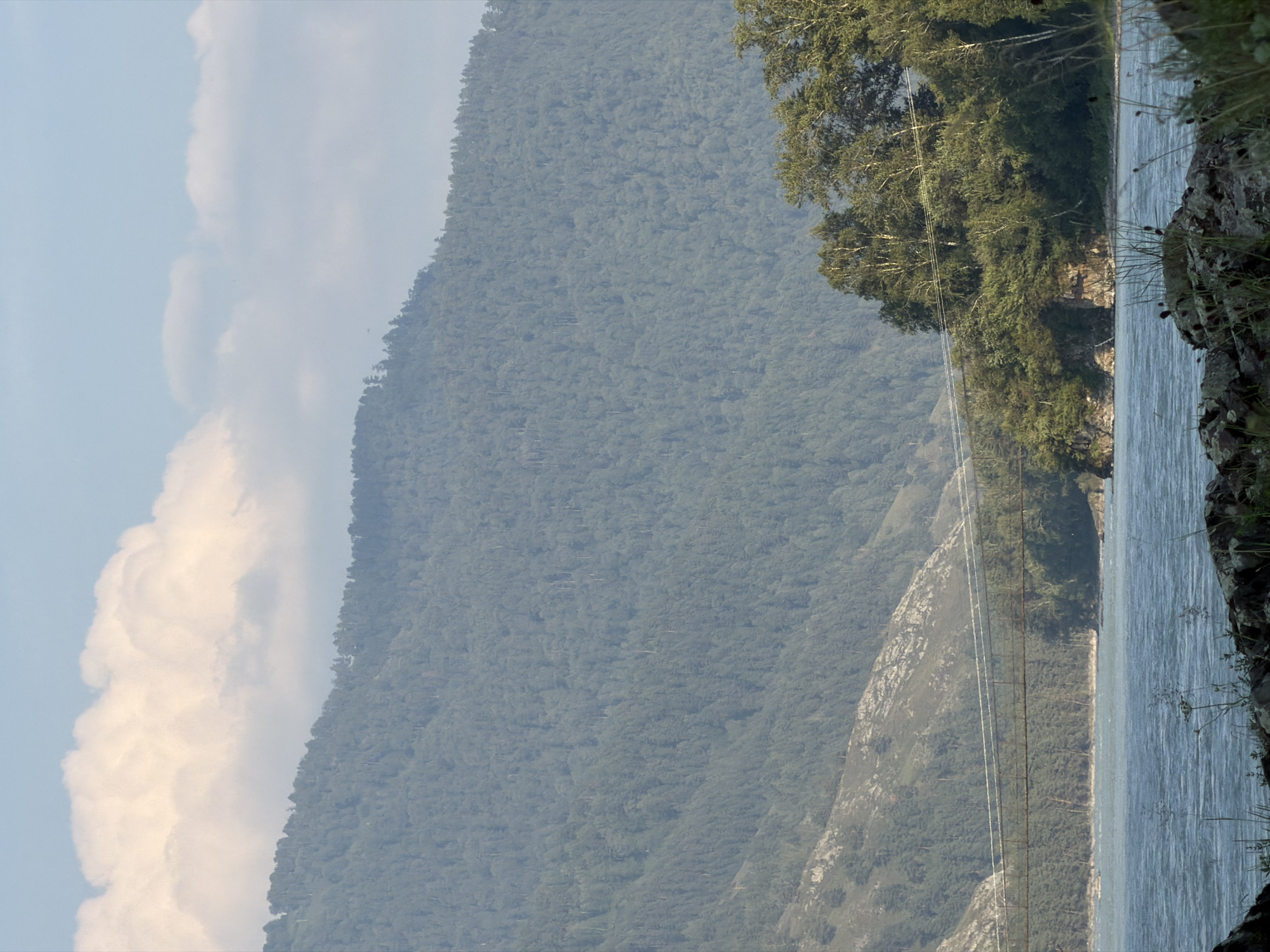 Anos, Russia. A wide, choppy river flows in the foreground, reflecting the sky. On the far bank, a dense forest covers a mountain slope. A thin suspension bridge spans the river, connecting the two banks. In the distance, another mountain, covered in trees, rises to meet a sky dotted with white clouds. The foreground features rocks and vegetation along the river's edge.