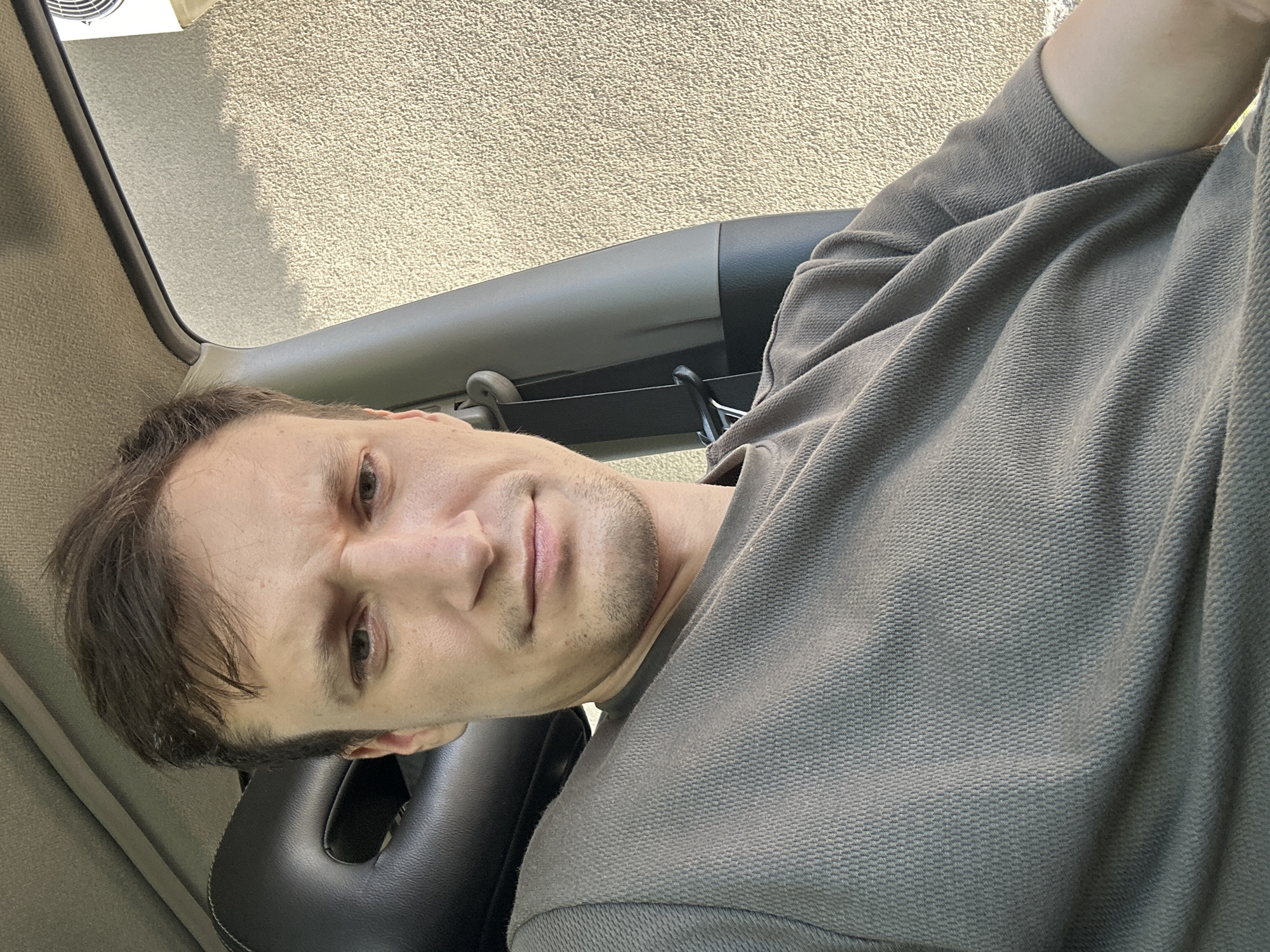 In Shatava, Ukraine, a selfie shows a man with short brown hair and light skin sitting in a car. He is wearing a dark grey t-shirt and his facial expression is neutral. The car interior is visible with a black headrest and a grey fabric lining on the ceiling. Outside the car, a beige textured wall is visible. The lighting suggests it is daytime.