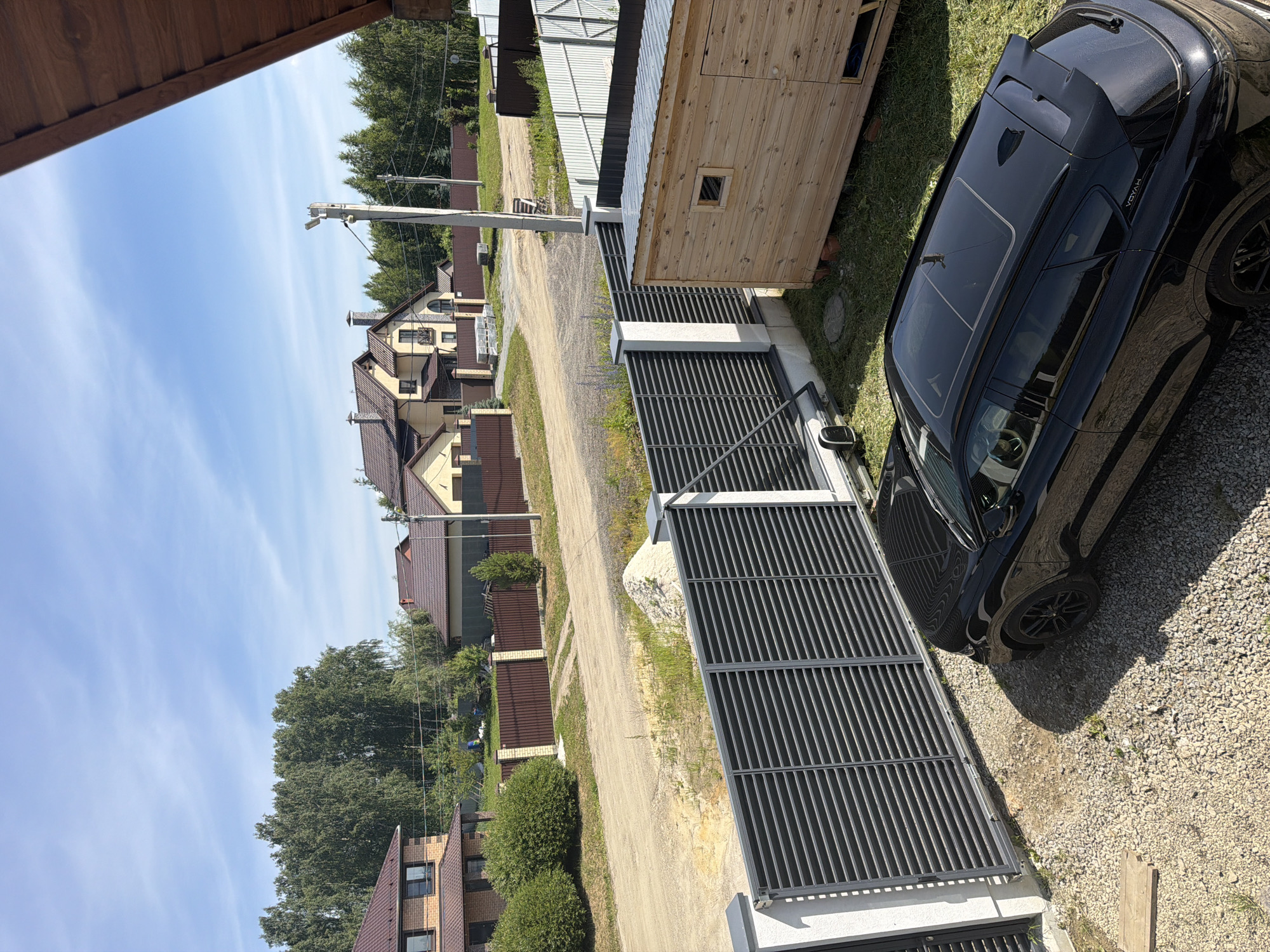 The image shows a residential street in Malinovka, Russia, on a sunny day with a blue sky and scattered clouds. In the foreground, a black SUV with a sunroof is parked on a gravel driveway next to a modern dark grey slatted fence. To the right of the fence is a small wooden outbuilding with a metal roof. In the background, there are several houses with red-tiled roofs, surrounded by trees and fences. A dirt road runs through the scene, and a utility pole with wires is visible. The overall impression is that of a suburban or semi-rural residential area. There are no people visible in the image.