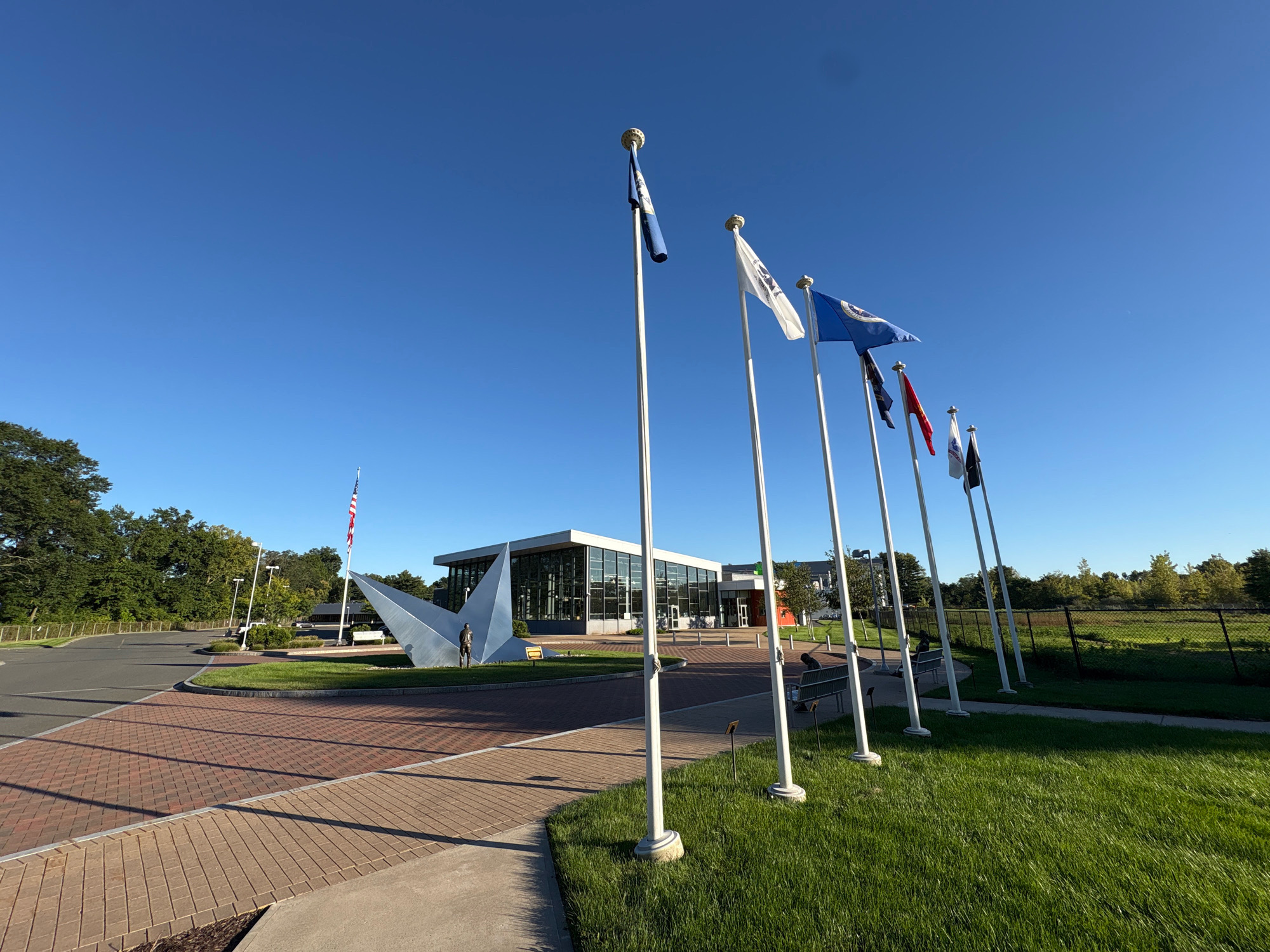 The image shows the exterior of a modern building with large glass windows, located in East Hartford, United States of America. In front of the building, a paved walkway leads to the entrance, with a circular area in the center featuring a large abstract metal sculpture and a statue of a person. A flagpole with an American flag stands to the left of the building. A row of flags on poles are positioned in front of the building, to the right of the walkway. The sky is clear blue, and the area is surrounded by green grass and trees. There are two benches visible on the grass.
