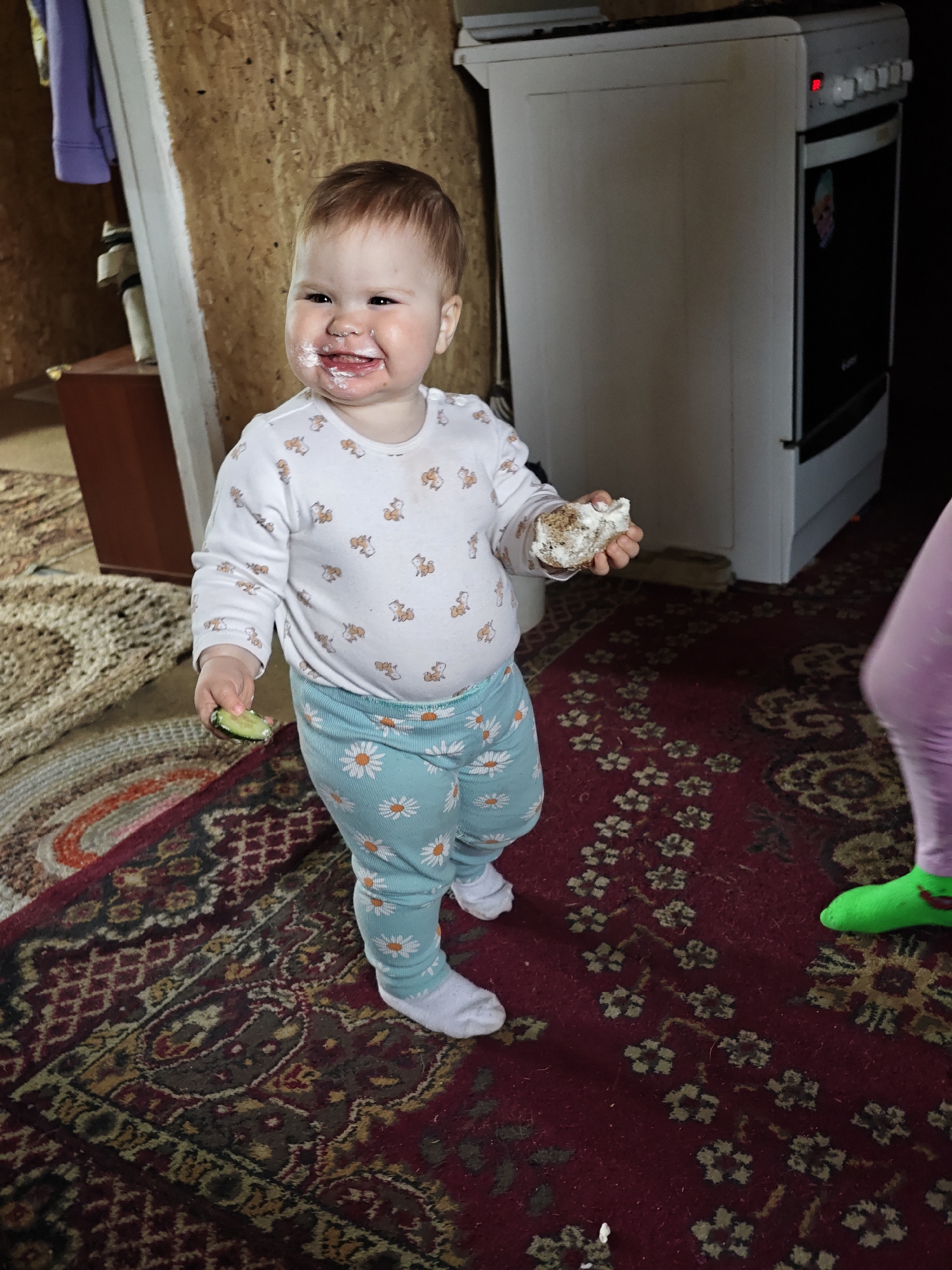 A toddler with fair skin and light brown hair, her face smeared with white creamy substance, is standing on a patterned rug indoors. She wears a white long-sleeved shirt with small animal patterns and light blue pants with a daisy print, along with white socks. In her right hand, she holds a chunk of cake covered in white cream, and in her left hand, she holds a slice of cucumber covered in cream. The rug is predominantly maroon with gold, brown, and black floral patterns. Behind the toddler is a white stove and a partially visible wall of plywood. A portion of another person’s leg wearing purple pants and a bright green slipper is visible on the right. The location is Malyi Bukor, Russia.