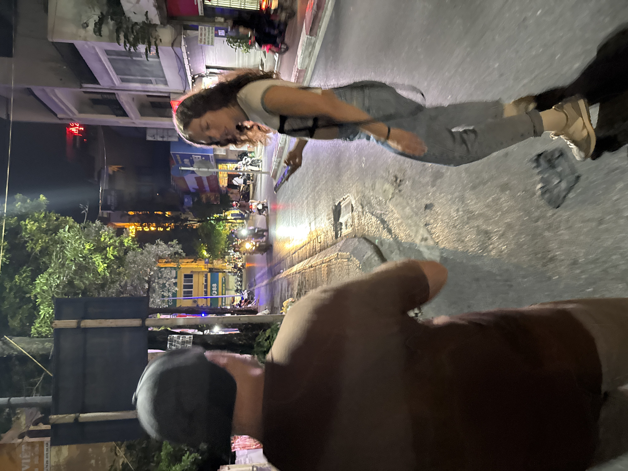 The media file captures a nighttime street scene in Hà Nội, Vietnam. The primary subjects are two people. In the foreground, a person wearing a brown shirt and a black helmet is partially visible, facing away from the camera. Their hand is raised, obscuring part of the view. The main subject, positioned centrally, is a young woman with long, dark hair. She is wearing a white t-shirt with a darker strap across her chest, and light-colored jeans. Her head is tilted down, and she appears to be looking at or interacting with a smartphone she holds in her right hand. Her expression is difficult to discern due to the angle and lighting, but her posture suggests engagement with the device.
The setting is a city street at night, with buildings lining one side of the road. The street is paved and appears to be well-lit by streetlights and lights from the buildings and vehicles. Several motorcycles are visible on the street, some parked and others in motion, indicating a common mode of transportation in this location. The atmosphere is lively, typical of a bustling urban environment after dark. The lighting casts reflections on the wet-looking pavement, suggesting recent rain or a generally humid climate.
Notable details include the vibrant signage on the buildings, some of which are illuminated with neon or LED lights. The presence of trees suggests a somewhat developed urban area with green spaces integrated into the cityscape. The time of day is clearly night, characterized by the artificial lighting and the darkness of the sky. The overall mood appears to be casual and everyday, with people going about their evening activities.
There is visible text on the buildings and potentially on signs or vehicles, but it is mostly illegible due to the image quality and distance. However, a red neon sign with Vietnamese characters is discernible on one of the buildings. The scene suggests a street in Vietnam, consistent with the provided location context of Hà Nội.