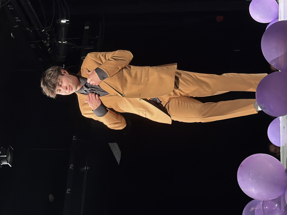An indoor scene depicts a young male standing center on what appears to be a stage in Palos Verdes Estates, United States. The individual wears a two-button tan suit jacket over a dark grey collared shirt and a dark tie, with matching tan trousers. White socks are visible above his footwear, which is obscured by balloons. His hands are positioned near his chest, adjusting the lapels or collar of the suit jacket. He has short, dark brown hair and is looking slightly down and to his right, with a subtle smile on his face. The background is predominantly dark, indicating a theater or performance space, with overhead stage lighting fixtures and cabling visible. White numbers, including "79," "78," "77," "24," and "25," are affixed to structures in the dark background. The foreground features several inflated, translucent purple balloons scattered across the stage floor.
