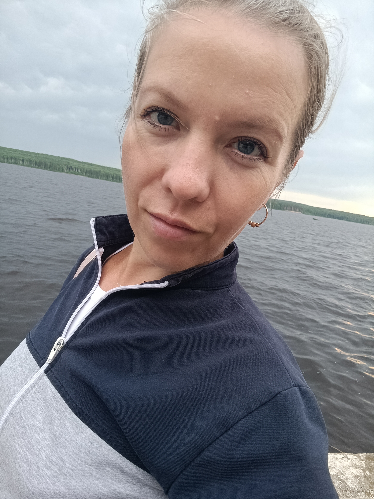 A woman is taking a selfie outdoors on a cloudy day by a body of water, likely a river or lake, with a tree-lined shore visible in the distance. She is wearing a navy and gray zippered jacket over a white shirt and a gold hoop earring. The water is choppy and a small boat is visible in the far distance. The woman's hair is pulled back and appears to be blonde or light brown. The overall scene suggests a casual outing. The location is likely near the Kama River in Chaikovsky, Russia, given the landscape and vegetation.