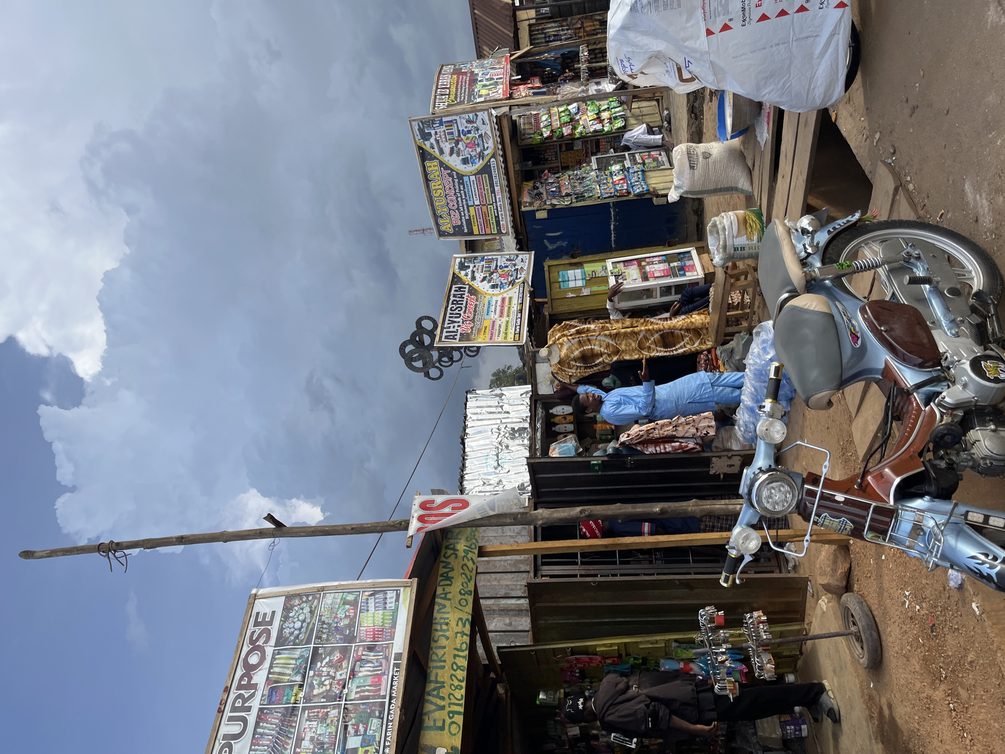 This image depicts a vibrant street market scene in Rafin Par, Nigeria. The foreground is dominated by a light blue and brown motorcycle, parked on a dusty, uneven ground. Behind the motorcycle, a bustling market unfolds with numerous stalls set up under a cloudy sky. Several signs are visible, advertising services such as printing, photocopying, typing, and binding, with names like "PURPOSE," "AL-YUSRAH Biz Concept," and "EVAFIRISHINA-DANSAR." The stalls are stocked with a variety of goods, including packaged items, what appear to be snacks or small goods, and larger bags of rice.
Several people are present in the market. One person in a blue outfit is seen behind a counter in one of the stalls, possibly attending to customers. Another person is partially visible wearing a dark jacket and hat, seemingly browsing or working at a stall. The overall atmosphere suggests a typical market day, with people engaged in commerce and daily activities. The weather appears overcast, indicating a possibility of rain or a generally cloudy day. The presence of multiple vendors and a variety of goods points to a well-established local market. The text on the signs provides specific contact information, including phone numbers and addresses, suggesting these are local businesses serving the community.