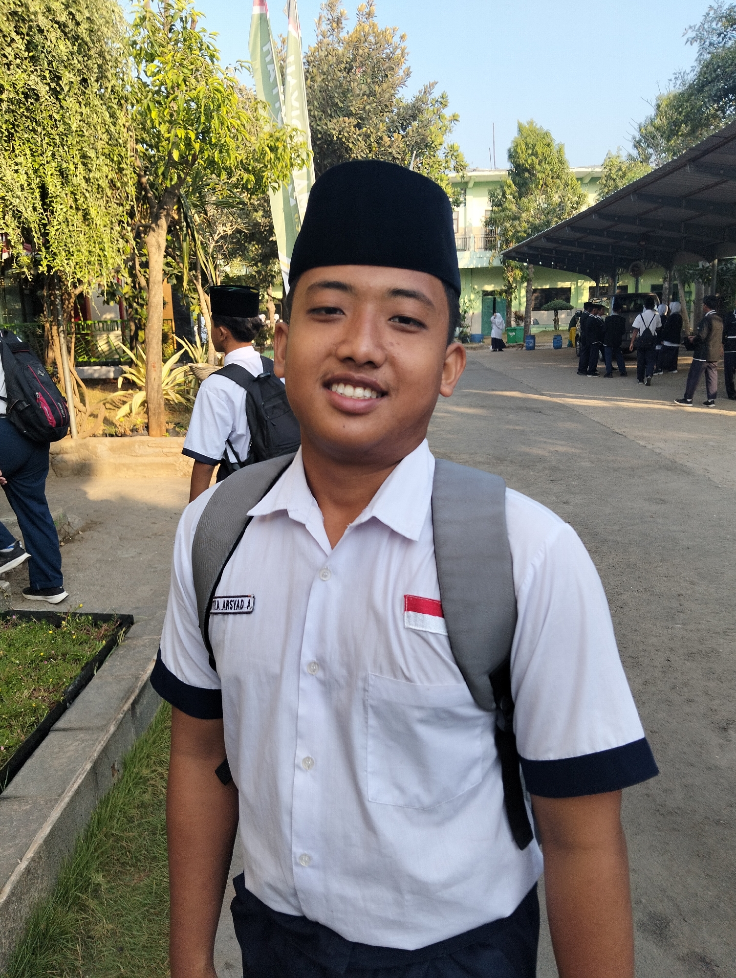 A teenage boy wearing a white school uniform shirt with dark trim, a black peci hat, and a grey backpack smiles at the camera. His shirt has a name tag that reads "YULIA ARSYAD A" and a small Indonesian flag patch on the left chest. He is standing outdoors on a dirt path next to a patch of grass and a concrete border. In the background, other students in similar uniforms are seen walking and conversing near a light green building with an awning. The environment suggests a school setting in Paciran, Indonesia, likely during the daytime, as indicated by the sunlight and clear sky.