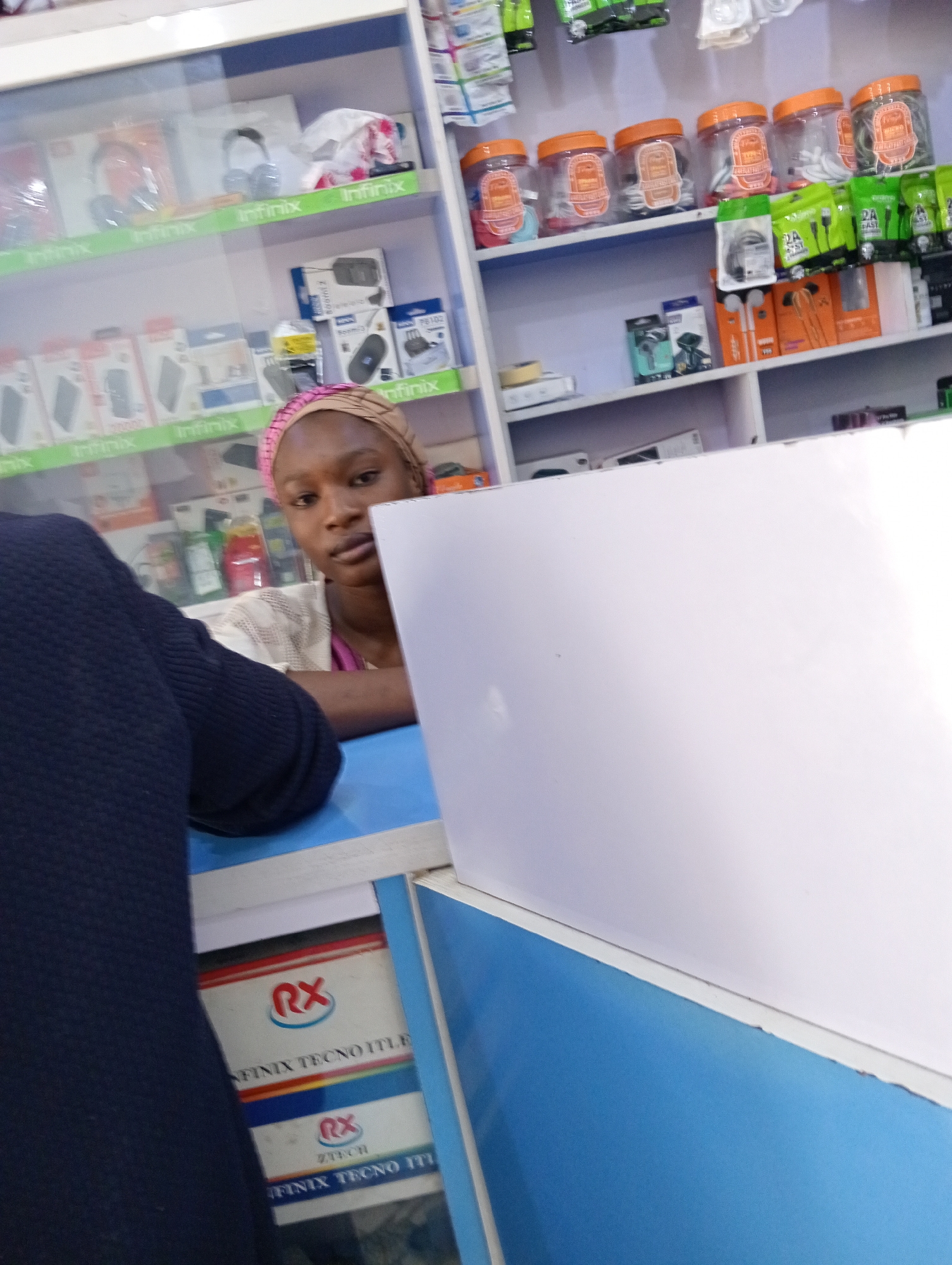 The image shows an indoor scene, likely inside a shop or store selling electronic accessories, possibly located in an urban area. A woman with a headscarf is standing behind a counter. Shelves behind her are stocked with various electronic accessories, including headphones, phone chargers, and cables, often packaged in branded boxes (e.g., "Infinix"). The items are neatly arranged, with some smaller accessories stored in clear plastic jars with orange lids. A person wearing a dark blue sweater is partially visible on the left side of the image, seemingly interacting with the woman behind the counter. A partition or display panel, predominantly white with blue edges, obscures a portion of the foreground. Lower down, an "RX" logo and the text "INFINIX TECNO ITLE" are visible on a box or sign.