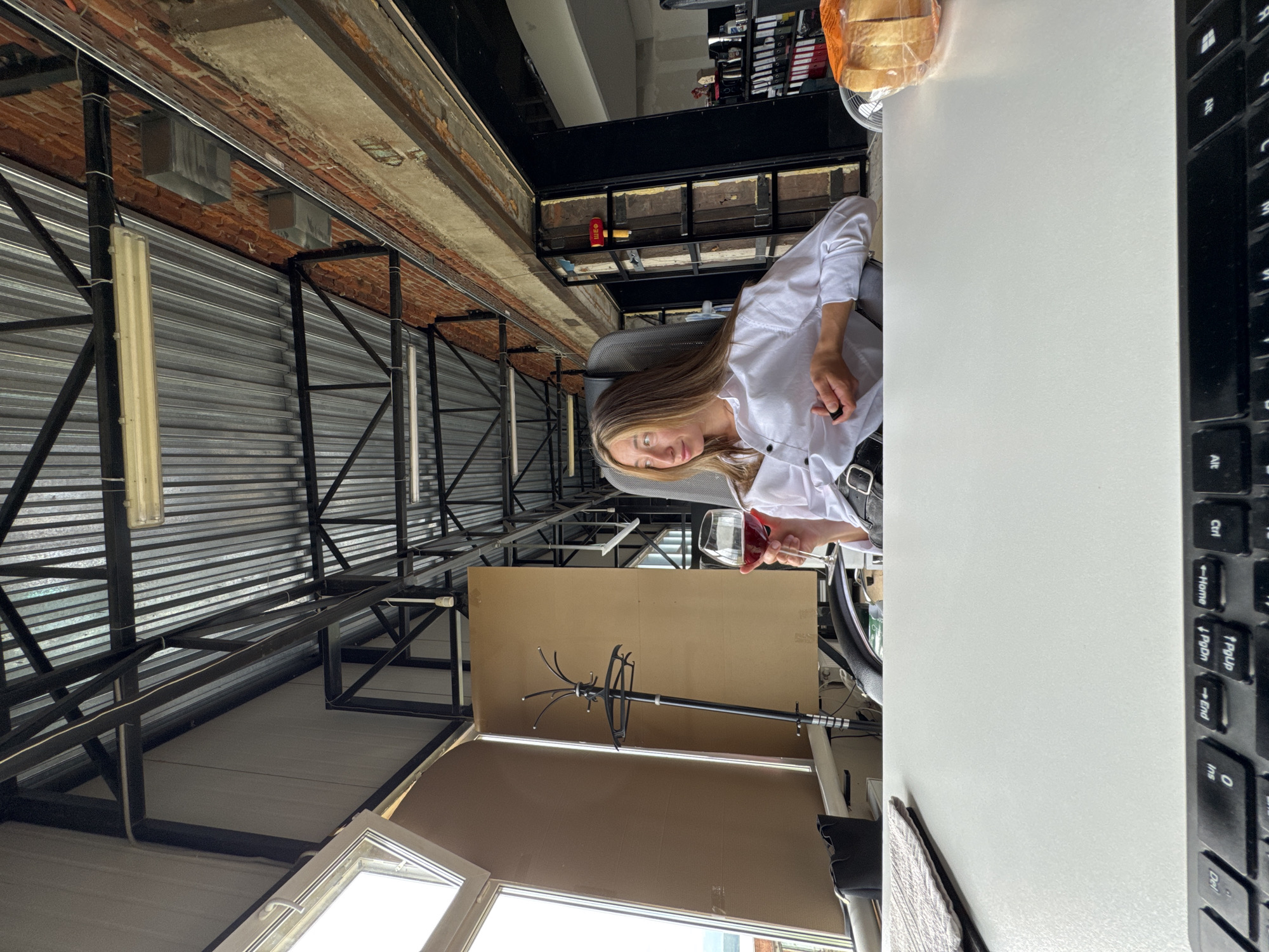 A woman with blonde hair is sitting in an office chair and holding a glass of red wine. She is wearing a white button-down shirt and black pants with a belt. The office has exposed brick walls, a metal ceiling, and large windows. There is a desk in front of her with a keyboard and some papers. The scene appears to be in an office space in Chaikovsky, Russia.