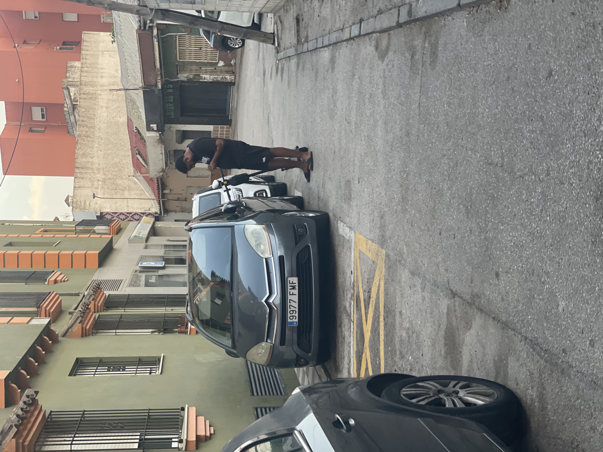 A medium shot captures an alleyway in La Línea de la Concepción, Spain, with a person standing next to an electric scooter and looking at their phone. The scene depicts a narrow street lined with buildings of varying architectural styles. On the left, a building with a green facade features windows with black security bars and terracotta-colored decorative elements. Opposite this, a more weathered building shows signs of age, with a corrugated metal roof.
A dark gray Citroën Picasso car is parked on the right side of the frame, its license plate clearly visible as "9977 FMF". A yellow criss-cross marking on the asphalt indicates a no-parking zone. The person, dressed in black shorts and a black t-shirt, is positioned between the parked car and a white electric scooter. Their posture suggests they are engaged with their mobile device. The ground is paved with asphalt, with a curb and some debris visible on the right side. The overall atmosphere is that of a typical urban street scene in a Mediterranean town.