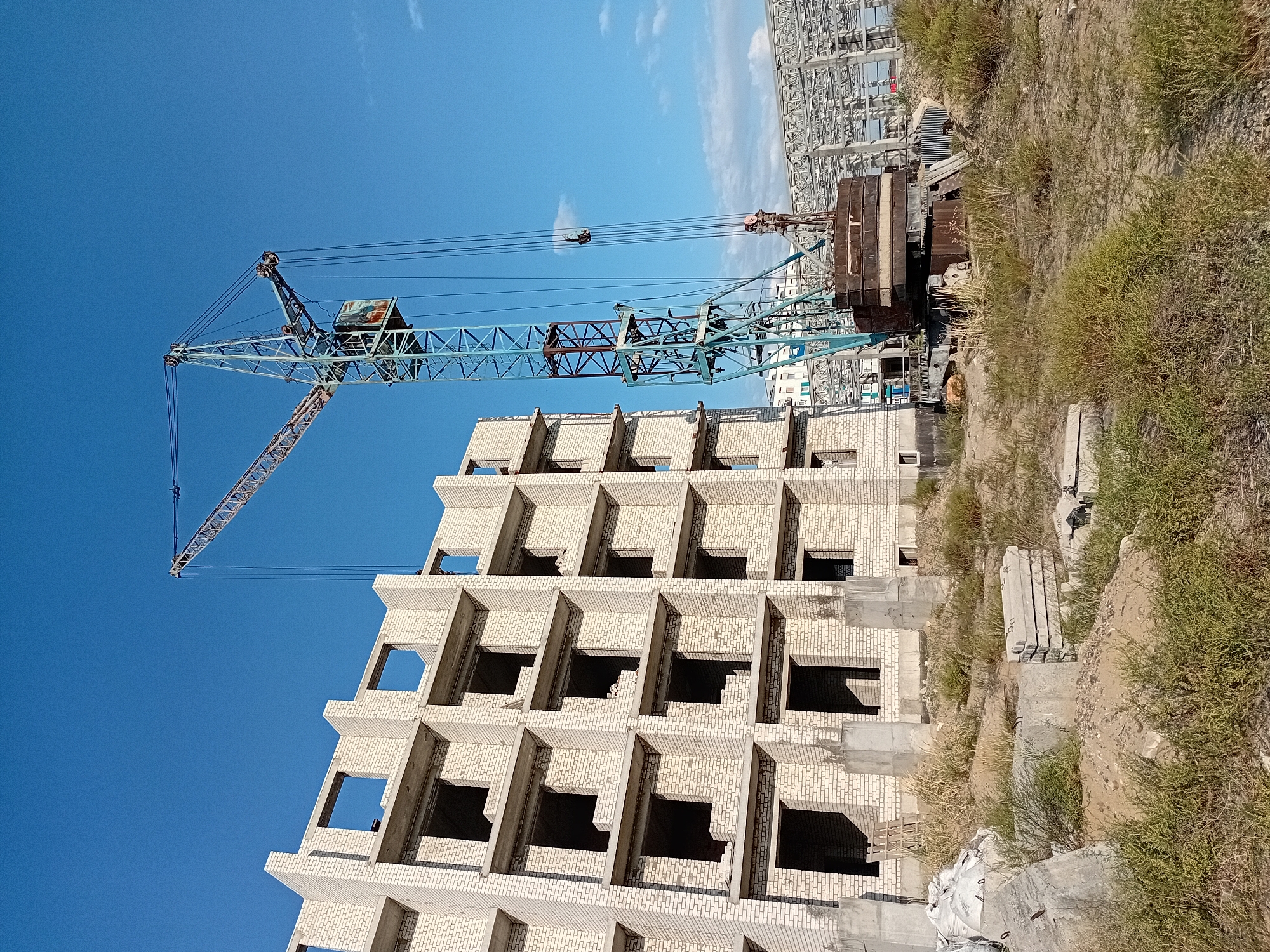 This is a vertical, eye-level, outdoor photograph of a construction site under a clear blue sky with a few wispy clouds. The primary focus is a multi-story building under construction, made of light-colored bricks with open rectangular window frames and concrete floors. To the right of the building stands a tall, blue and brown tower crane with visible cables and a metal operator's cabin. The base of the crane is adorned with large, circular metal counterweights. The ground in front of the building and crane is covered with dry grass, sand, and various construction materials, including concrete slabs, bags, and debris. In the background, a large, unfinished structure with a metal frame is visible. The overall scene suggests an active construction project in the city of Semey, Kazakhstan. There are no people visible in the photograph.