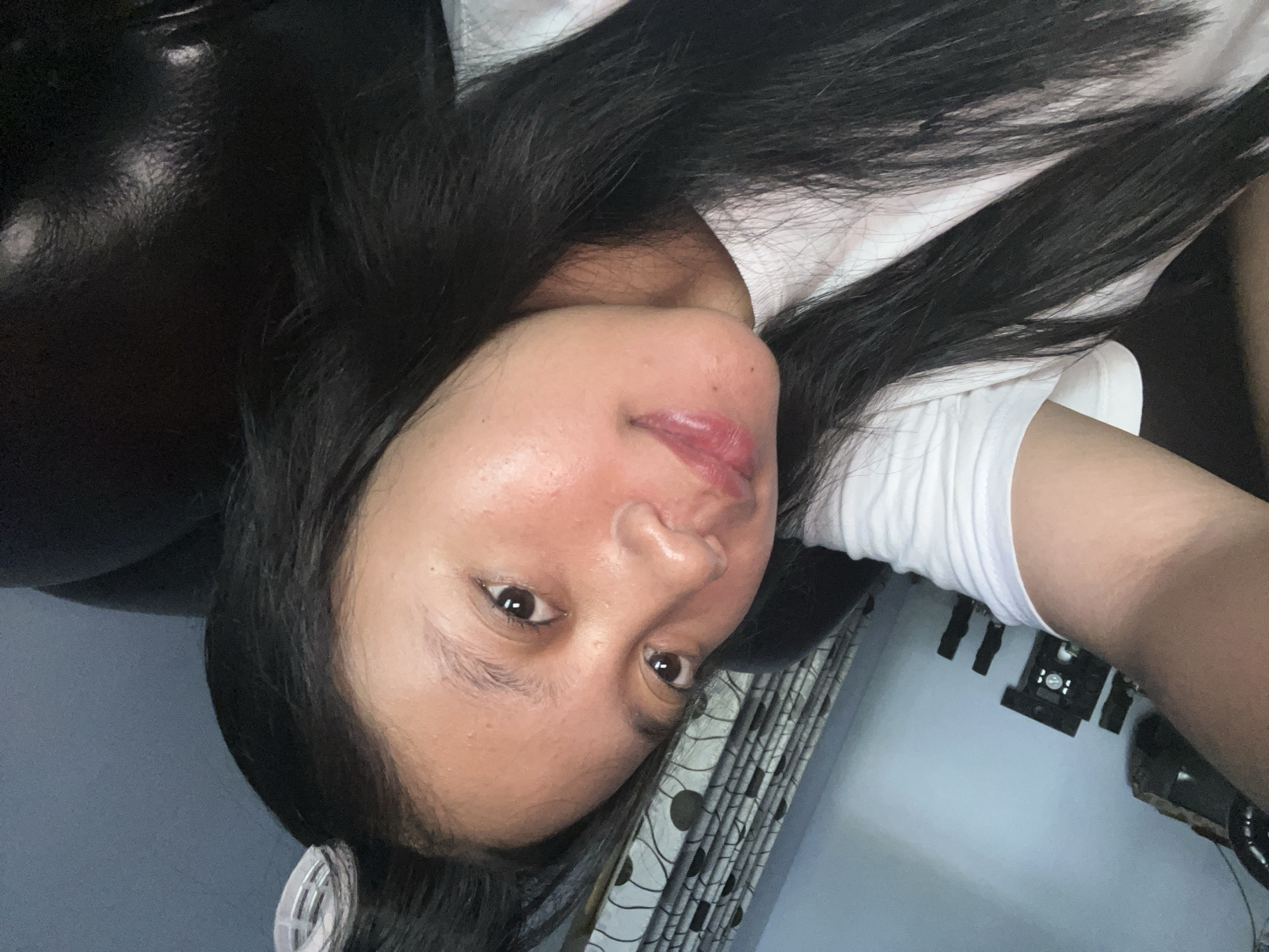 A young woman with black hair and dark eyes is taking a selfie. She is wearing a white t-shirt and has a hair roller in her hair. She appears to be indoors, possibly in a bedroom or living room, as suggested by the presence of a curtain and furniture in the background. The lighting is dim, indicating it might be evening or nighttime. There are no other people visible in the image. The scene is a personal, candid moment captured by the woman herself.