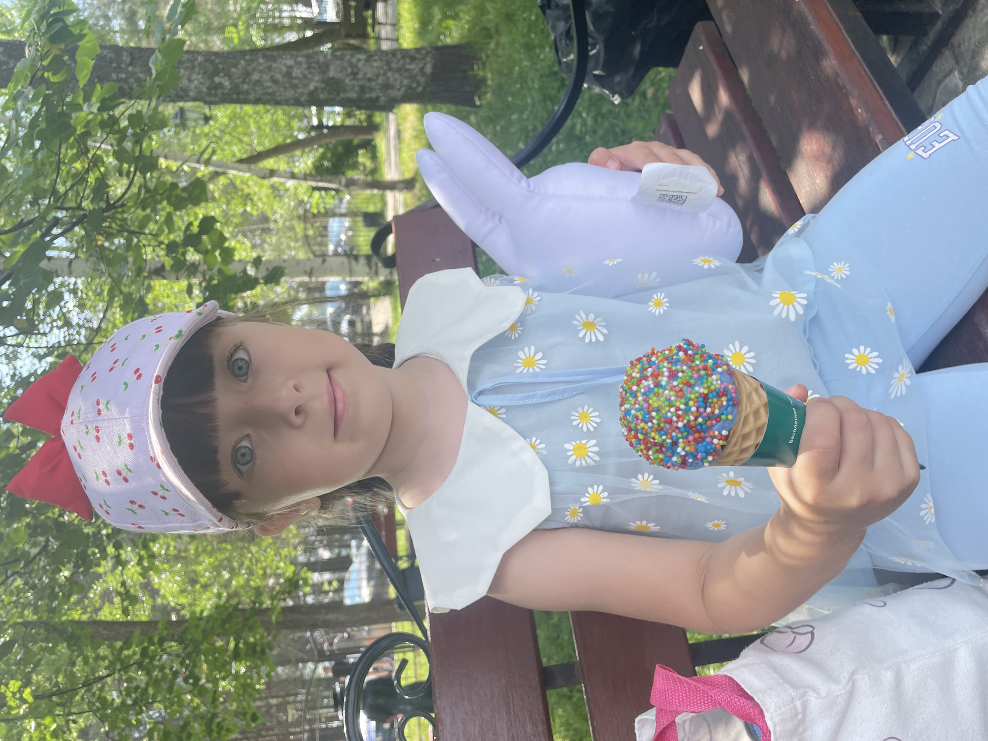 A young girl, with blue eyes and dark bangs, sits on a park bench, holding an ice cream cone topped with colorful sprinkles and a white stuffed bunny. She wears a light blue dress with a white collar adorned with daisies and a white baseball cap with a cherry pattern and a red bow. The park setting features trees and a clear sky. The girl is likely in Chaikovsky, Russia, given the context of previous images from this location.