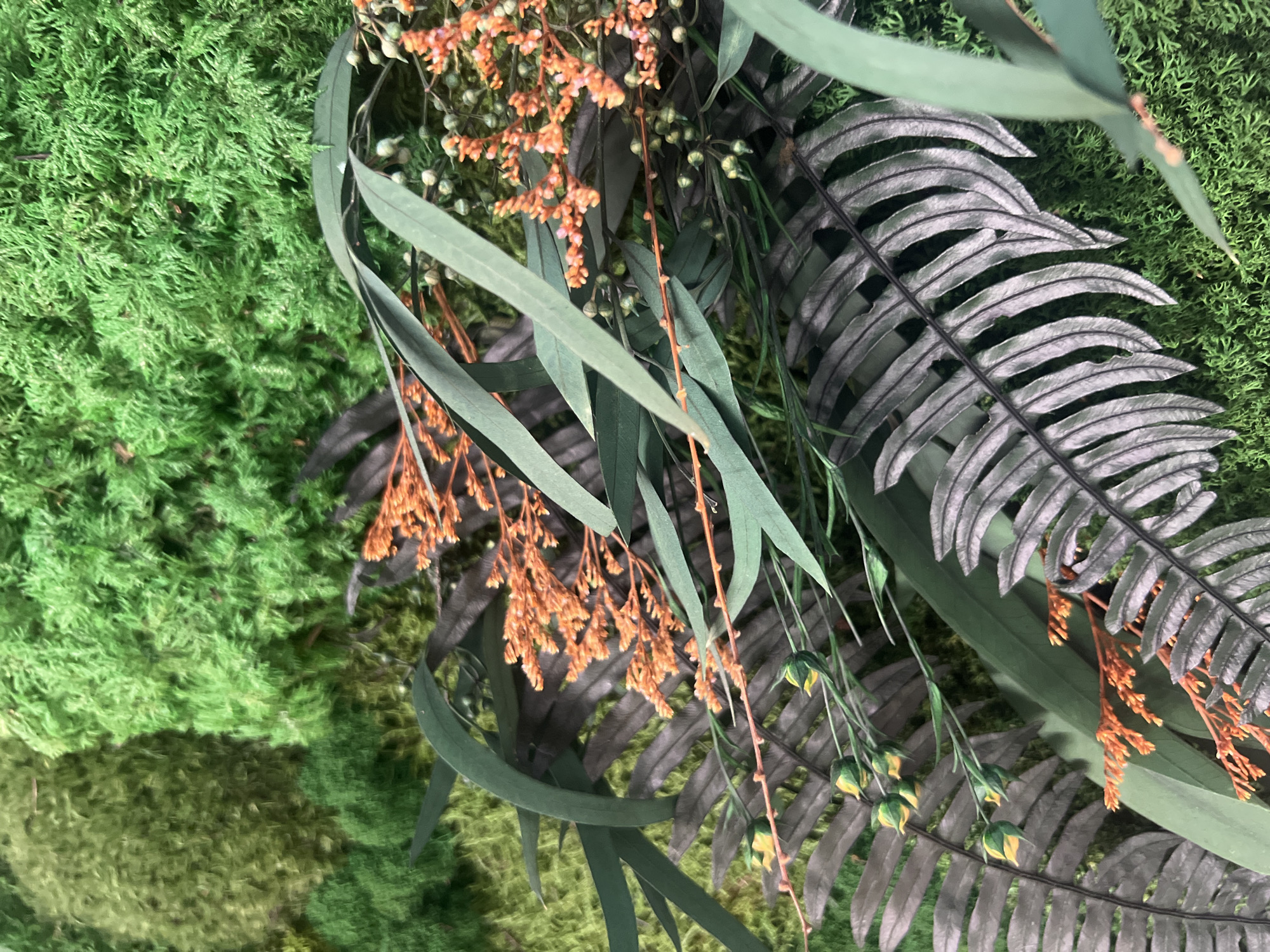 This image displays a close-up of a decorative arrangement of faux greenery and flowers, likely a floral installation or a part of a larger botanical display. The composition features a vibrant, textured green moss as the background, interspersed with various artificial plants. Prominently featured are long, slender green leaves, resembling eucalyptus, and dark, gray-green fern fronds with finely dissected leaflets. Delicate sprays of small, orange-red flowers and buds add a contrasting pop of color and texture. Some green buds, possibly from a different floral element, are also visible. The lighting is even, suggesting an indoor setting or diffused daylight. There is no discernible text within the image, and no people or specific activities are depicted. The overall impression is one of lush, naturalistic, but artificial, botanical beauty. The context of "Minsk, Russia" doesn't provide specific visual cues within the image itself, as the arrangement is a common decorative element.