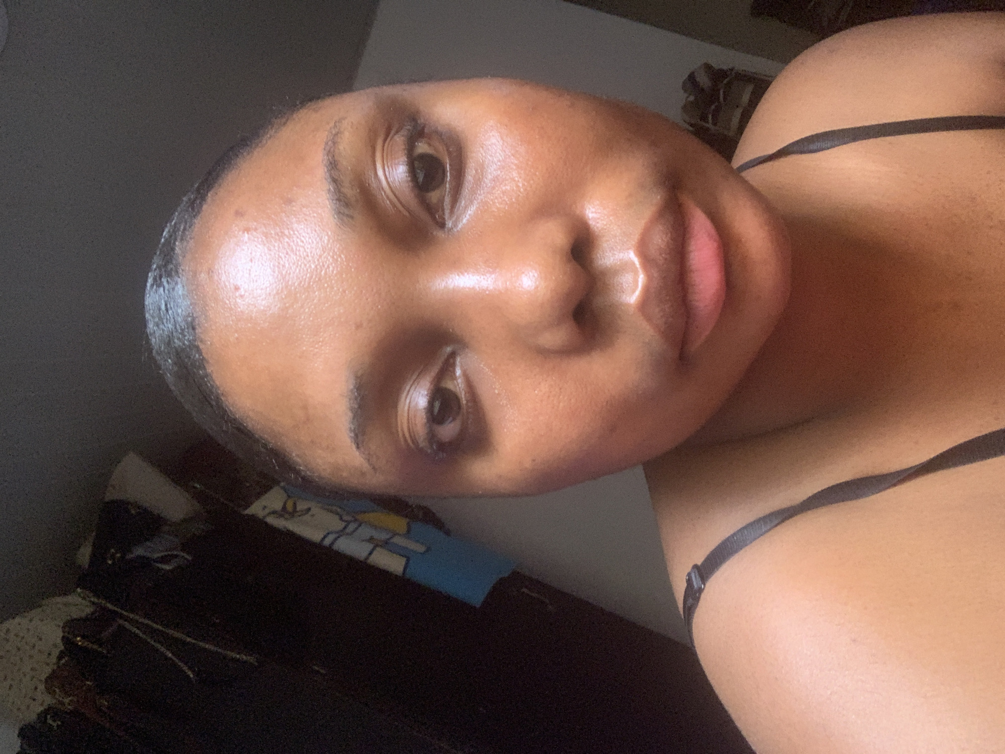 A close-up selfie shows a young woman with brown skin. She has smooth skin, short dark hair, and full lips. A thin black strap crosses her right shoulder. The background is dark and cluttered, with a cabinet and bedding visible. The location is unspecified, and there is no indication it is in Abuja, Nigeria.