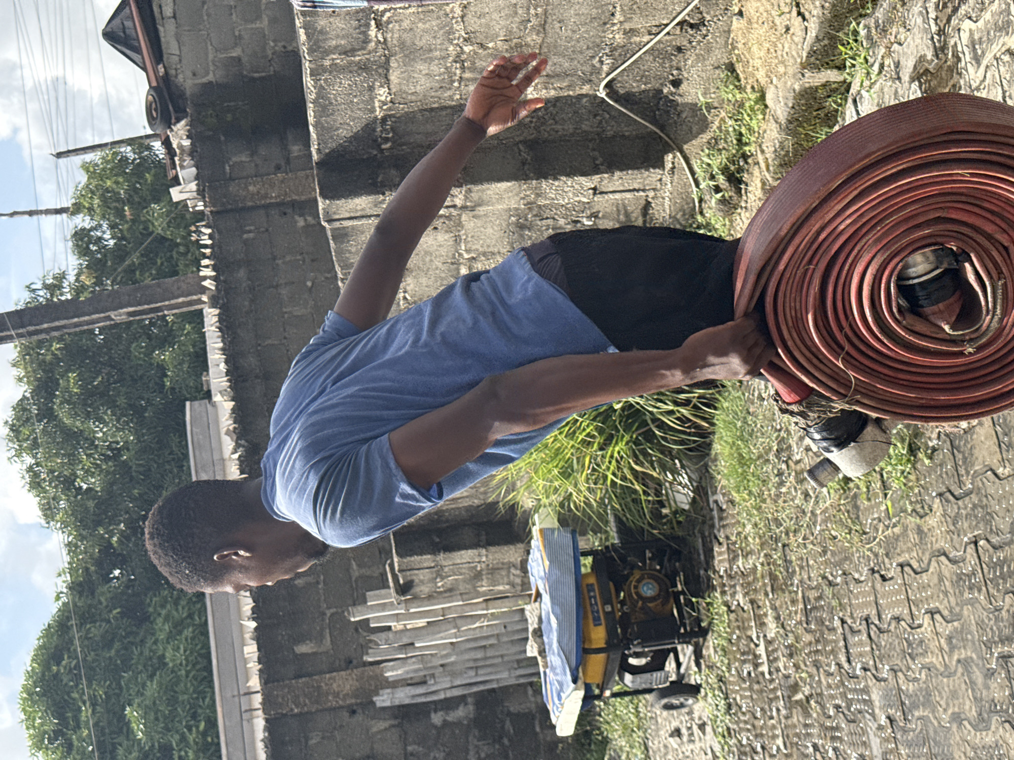 A man is holding a coiled red fire hose. He is wearing a blue shirt and black pants. He is standing on a paved surface, and a yellow generator is nearby. In the background is a concrete block wall and trees. The location is in Ado, Nigeria.