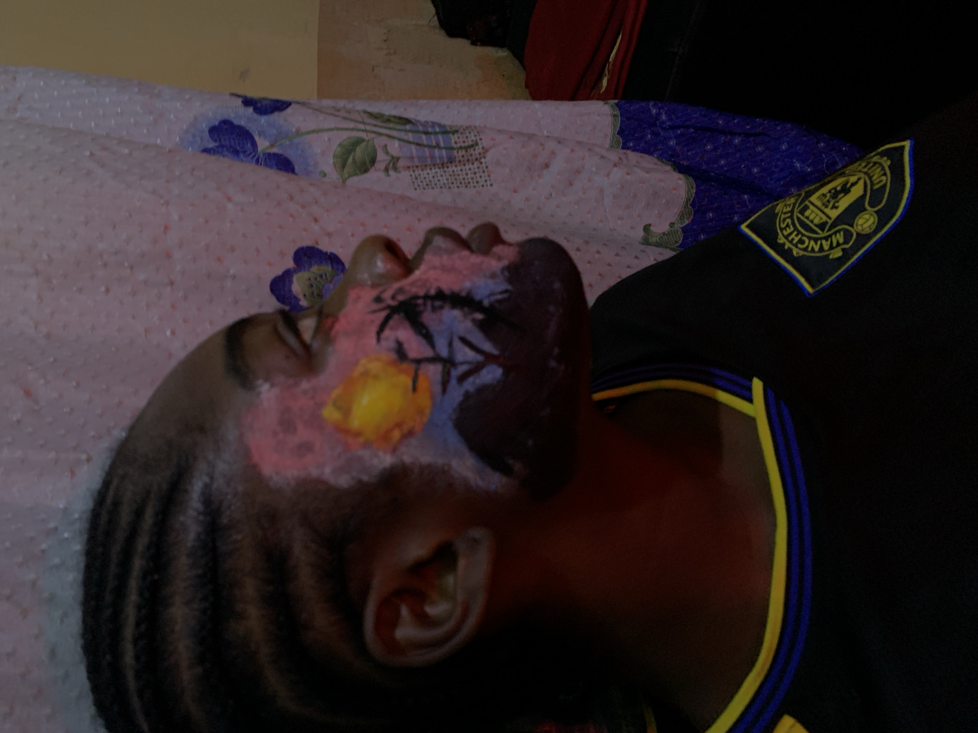 The image shows a close-up of a person's face with elaborate face paint. The face paint features a mix of colors, including pink, yellow, blue, and black, with black designs resembling branches or lightning. The person has braided hair on the side of their head and is wearing a black shirt with a yellow and blue trim around the collar. On the shirt's shoulder, there is a patch with a crest, clearly visible as the Manchester United Football Club logo, complete with the text "MANCHESTER UNITED." The background appears to be a bed with a patterned duvet. The lighting suggests it is indoors, likely during the day. The overall impression is that of a person dressed up or participating in an event where face painting is involved, possibly related to supporting Manchester United. The location context is Rafin Par, Nigeria.