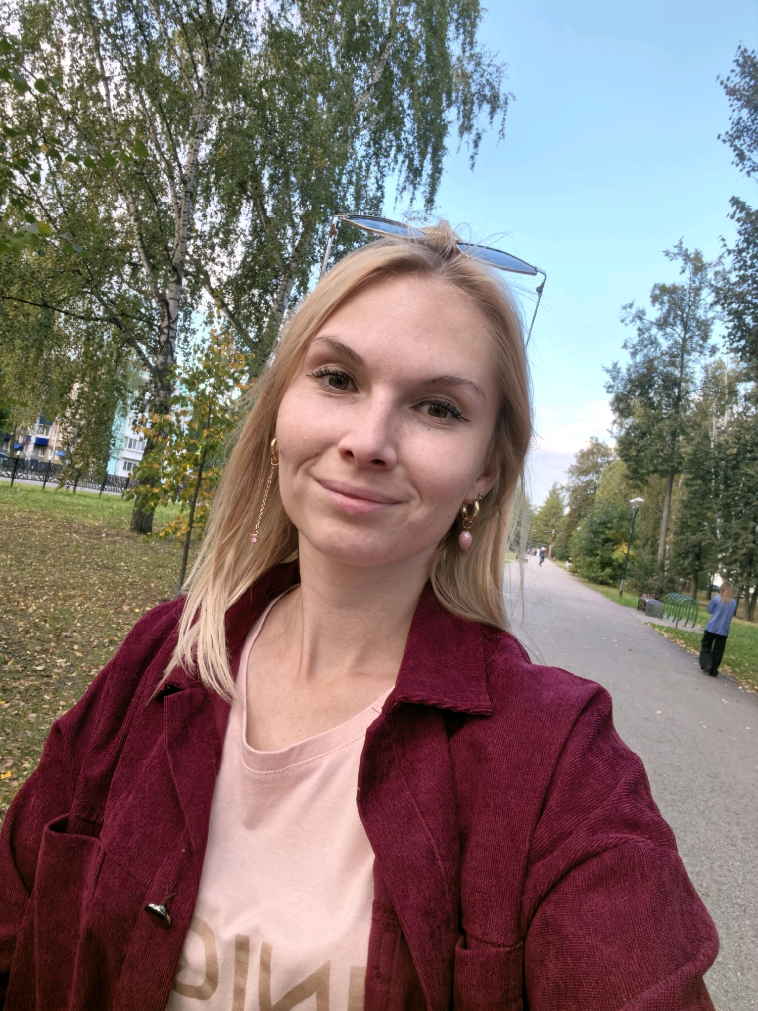 In Sterlitamak, Russia, a blonde woman with fair skin and light makeup takes a selfie in a park. She wears a maroon corduroy jacket over a light pink t-shirt with a gold graphic. A pair of sunglasses rests on her head, and a gold earring with a pink stone hangs from her left ear. The park features a paved path with trees and fallen leaves on either side. In the background, a person in a blue jacket walks away from the camera. A building and more trees are visible further in the distance. The sky is clear and blue.