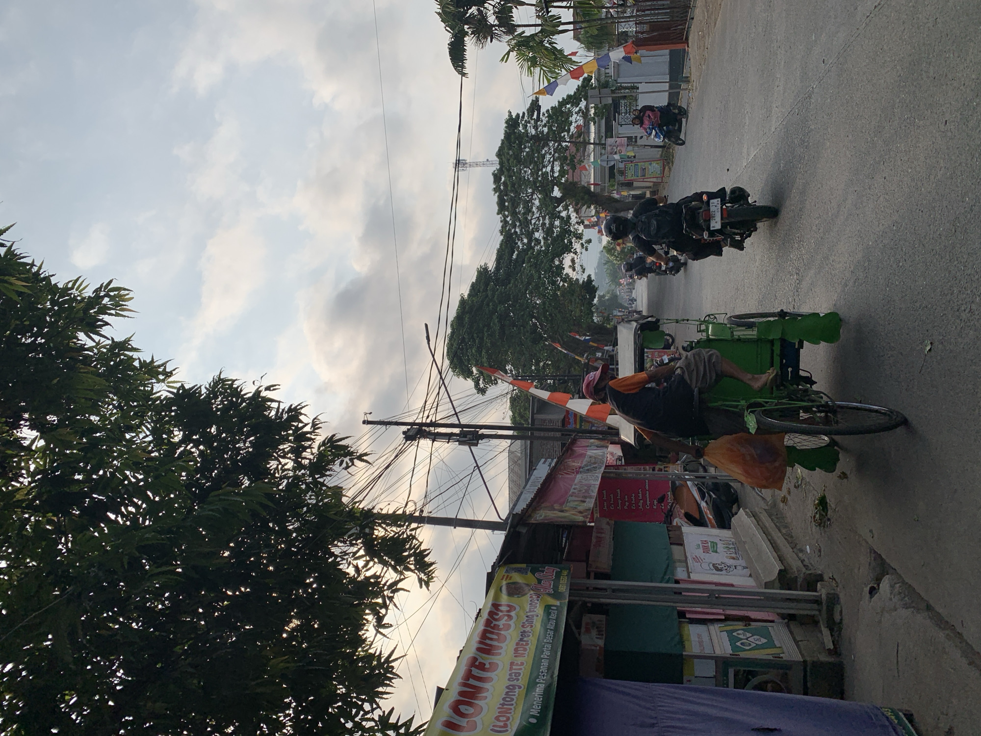 The image depicts a street scene in Majenang, Indonesia, likely during the late afternoon given the soft lighting and cloud cover. On the left side of the road, a shop with a banner that reads "LONTE NDESO" is visible, indicating it sells food, possibly "Lontong Sate Nderek Sing Kudus." The shop appears to be open, with offerings displayed.
Dominating the foreground is a green pedicab, with a person driving it and an orange plastic bag containing items in the passenger area. The driver is wearing a red hat and an orange shirt with a black vest. The pedicab is moving along the road.
Further down the street, several motorcycles are in motion. One motorcycle in the mid-ground has a rider with a black backpack, and another motorcycle in the distance has two people on it, one of whom is wearing a pink jacket. Various flags, including Indonesian flags and colorful pennants, are displayed along the roadside, adding to the local ambiance.
The environment includes trees with dense foliage, utility poles with numerous wires, and buildings lining the street. The sky is partly cloudy, with soft light filtering through. The road surface is asphalt, and there is a sidewalk on the left side of the street where the shop is located. The overall scene conveys a sense of everyday life and local commerce in a bustling Indonesian town.