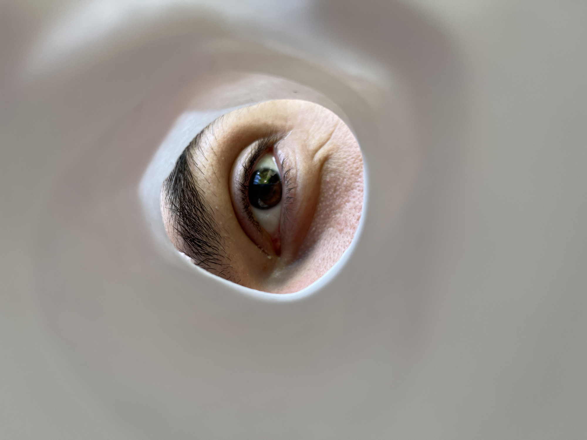 An extreme close-up shot shows a human eye looking through a circular opening in a white, amorphous object. The eye is brown, with visible eyelashes and a slightly wrinkled eyelid. The surrounding white object is out of focus and creates a vignetting effect. The lighting suggests the scene is illuminated by natural light. The image does not contain any identifiable landmarks or features that would indicate its location in Sofia, Bulgaria.