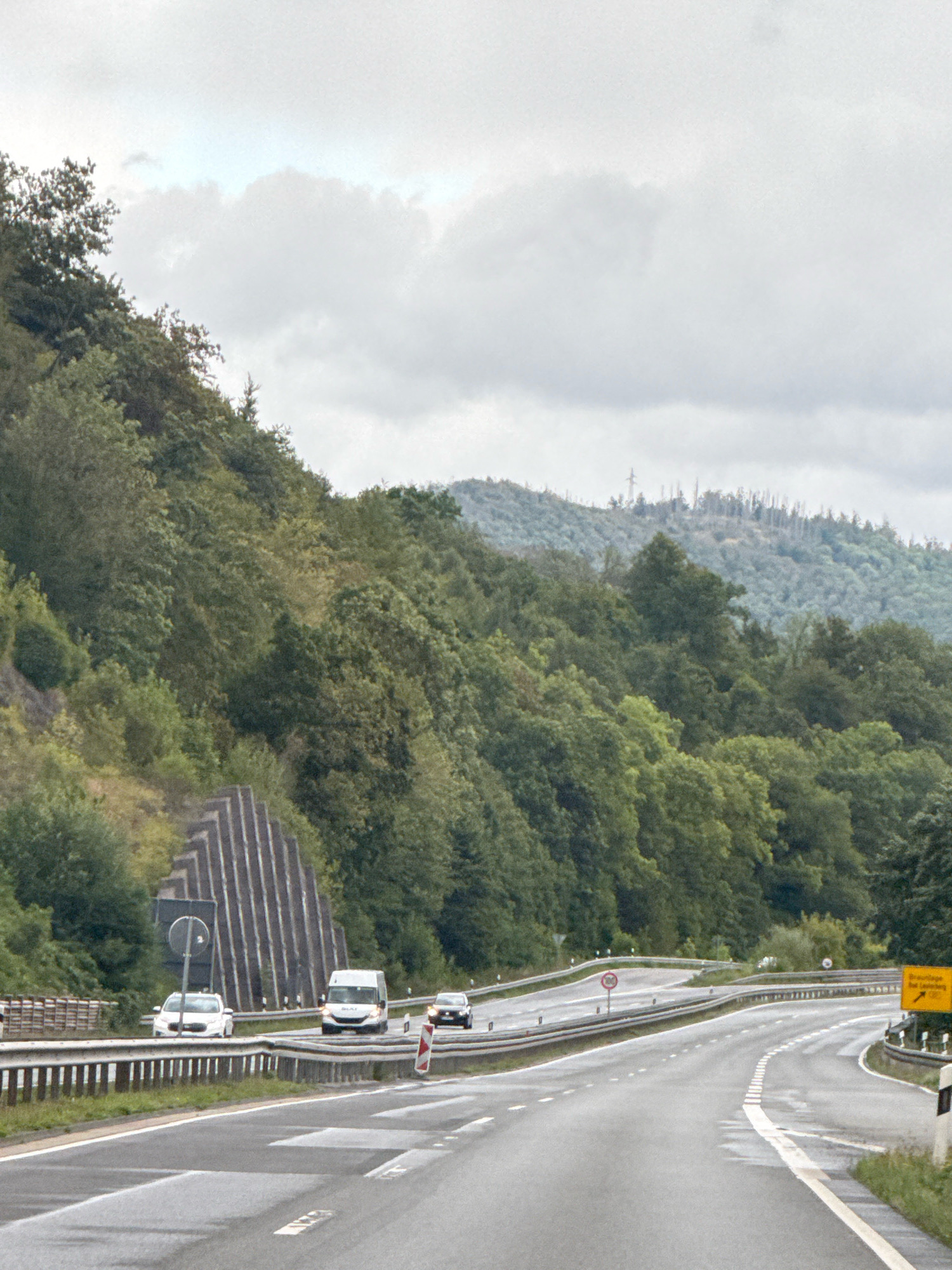 This image captures a view of a highway in or near Bad Lauterberg im Harz, Germany, under a cloudy sky. The highway has multiple lanes and is bordered by guardrails. Several vehicles, including cars and a white van, are visible on the road. To the left, a steep, tree-covered hillside rises, with a section of textured concrete retaining wall. In the distance, more rolling hills covered in dense forest are visible, with some areas appearing to have dead trees. A yellow road sign with German text is present on the right side of the highway. The road surface appears wet, suggesting recent rain.
