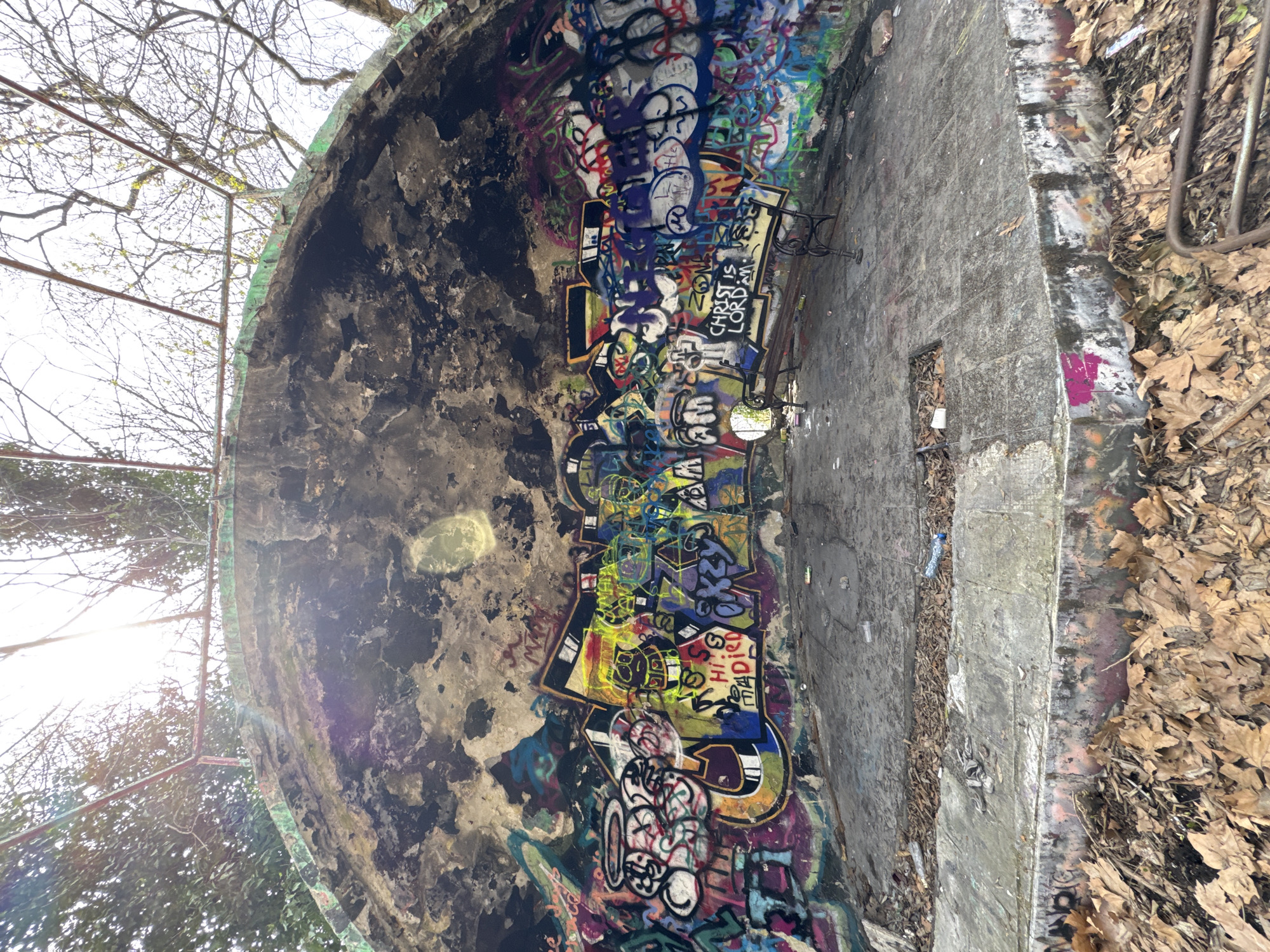 The image shows the interior of a concrete structure, possibly a bunker or underground reservoir, covered in extensive graffiti. The structure is circular or semi-circular in shape, with a flat floor and curved walls leading upwards. The concrete surfaces are weathered and stained. The graffiti is multicolored and dense, obscuring much of the original concrete.
To the right of the frame, the edge of the structure leads to a ground covered in dead leaves. A metal structure is visible, perhaps a railing or fence. Through an opening on the top of the structure, trees and sky are visible.
The location, Varna, Bulgaria, is not visually identifiable within the scene.