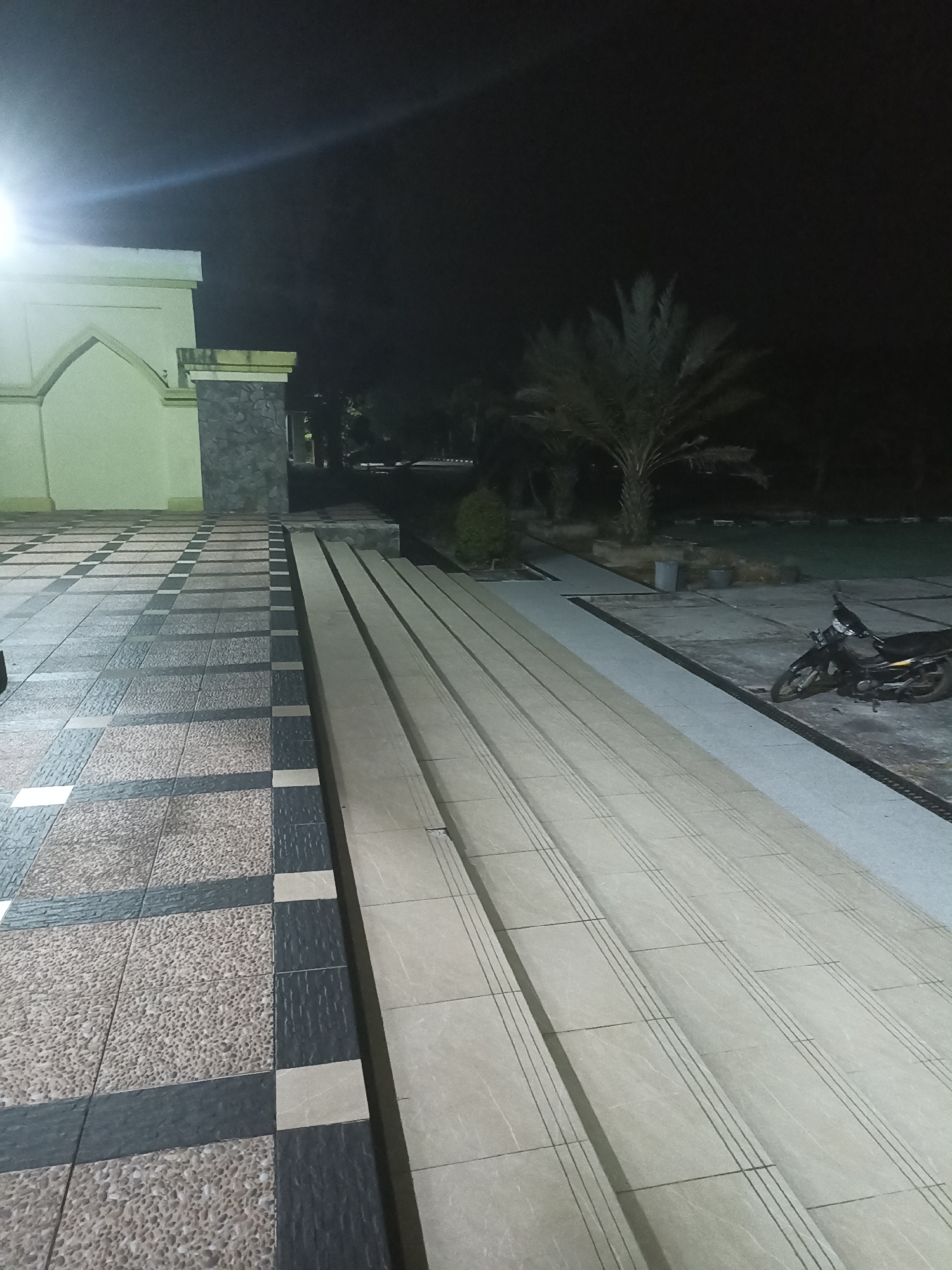 At night in Sungai Mempura, Indonesia, a wide tiled walkway leads to a set of stairs. The walkway is laid with alternating patterns of light brown speckled tiles and dark grey rectangular tiles, interspersed with cream-colored square tiles. To the left, a light-colored building with arched window frames is visible, and to the right, a motorcycle is parked on a concrete surface. Beyond the motorcycle, a palm tree stands against the dark night sky. The stairs are made of light beige tiles with subtle horizontal grooves. The overall scene is illuminated by artificial light, creating shadows and highlighting the textures of the various surfaces.