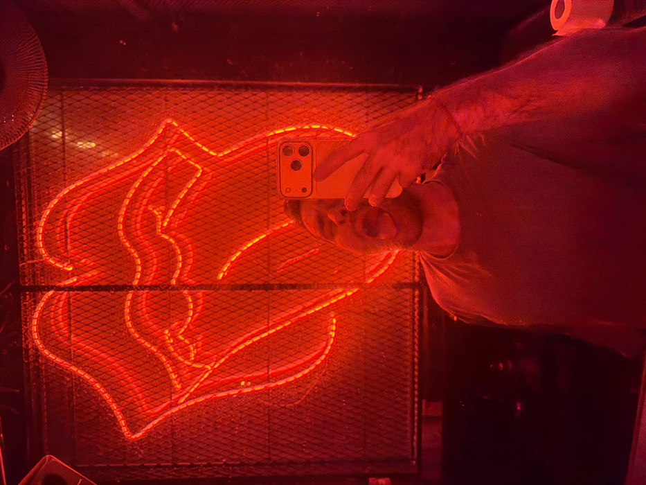 A person is taking a selfie in a mirror with a yellow smartphone. The mirror reflects a red neon light in the shape of a stylized flame or abstract design, mounted on a metal mesh. The entire scene is bathed in a strong red light, casting red hues onto the person, the phone, and the surroundings. A roll of toilet paper is visible in the upper right corner. Part of an electric fan is visible to the left.
