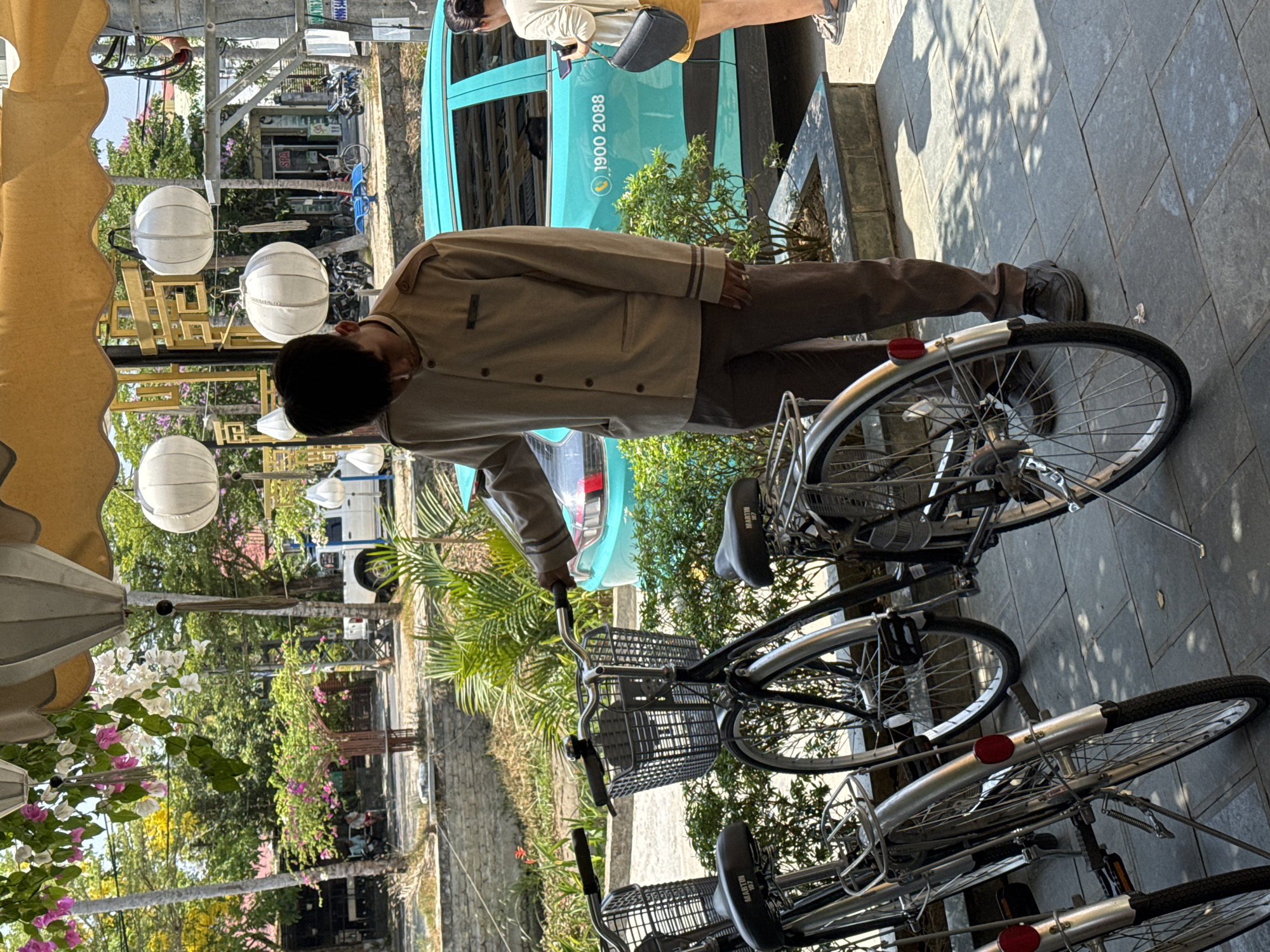 In Hội An Ward, Vietnam, a person in a light brown uniform is standing near a row of parked bicycles. The bicycles are mostly black and silver with wire baskets in the front. A turquoise vehicle, with the phone number "1900 2088" displayed, is parked in the background. Street lanterns hang in the scene, and trees and plants fill the surrounding environment.