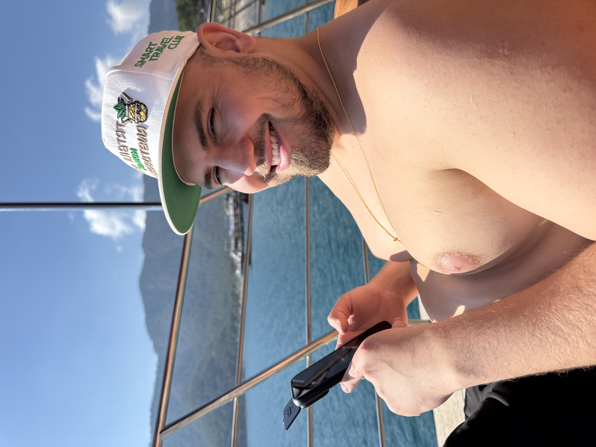 A young man, shirtless and with a beard, is smiling broadly towards the camera, suggesting a cheerful and relaxed mood. He is wearing a white baseball cap with a green brim, adorned with a pineapple logo and the text "SMART TRAVEL CLUB" in English, and "SMART ТУРИЗМ ПУТЕШЕСТВИЯ" (Smart Tourism Travel) in Russian. A thin gold chain is visible around his neck.
He is holding a black, compact device, possibly a portable mini-fan or a utility tool, in his hands. The setting is outdoors, likely on a boat or a dock, with a metal railing separating him from the water below. The background features clear blue water with ripples, suggesting motion, and distant, hazy mountains under a bright blue sky with a few white clouds. The strong sunlight indicates it is daytime. The man's skin appears healthy and possibly tanned. The overall scene evokes a vacation or leisure activity in a scenic coastal or lakeside location like Kumluca, Turkey.